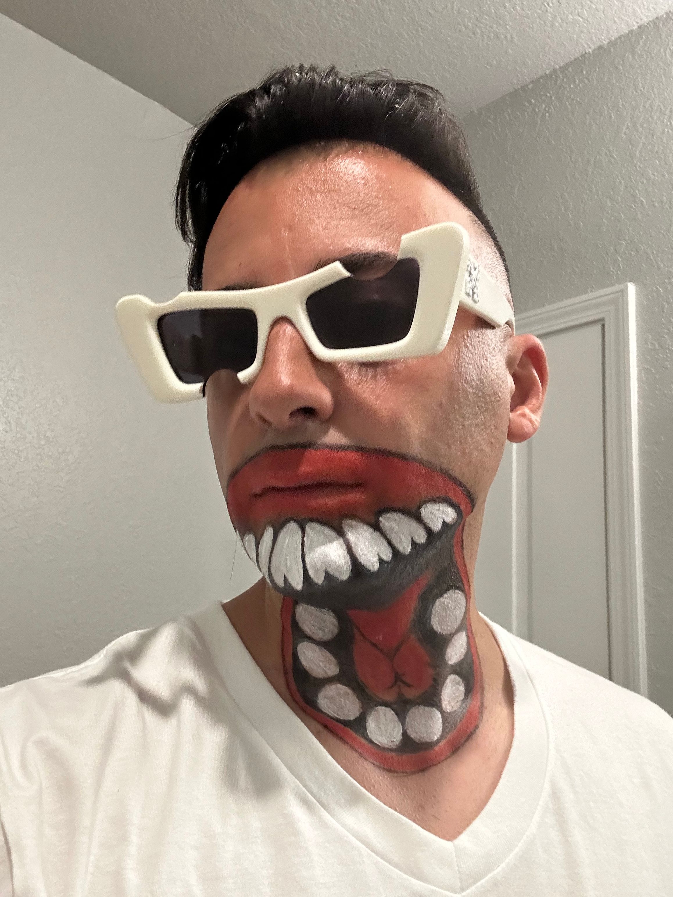 Person wearing sunglasses with a painted open mouth on neck, showing teeth and tongue, in a white shirt, against a plain wall.