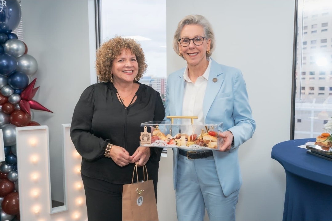 Two women smiling, one holding a tray with food, standing indoors near windows, decorated with balloons and a table.