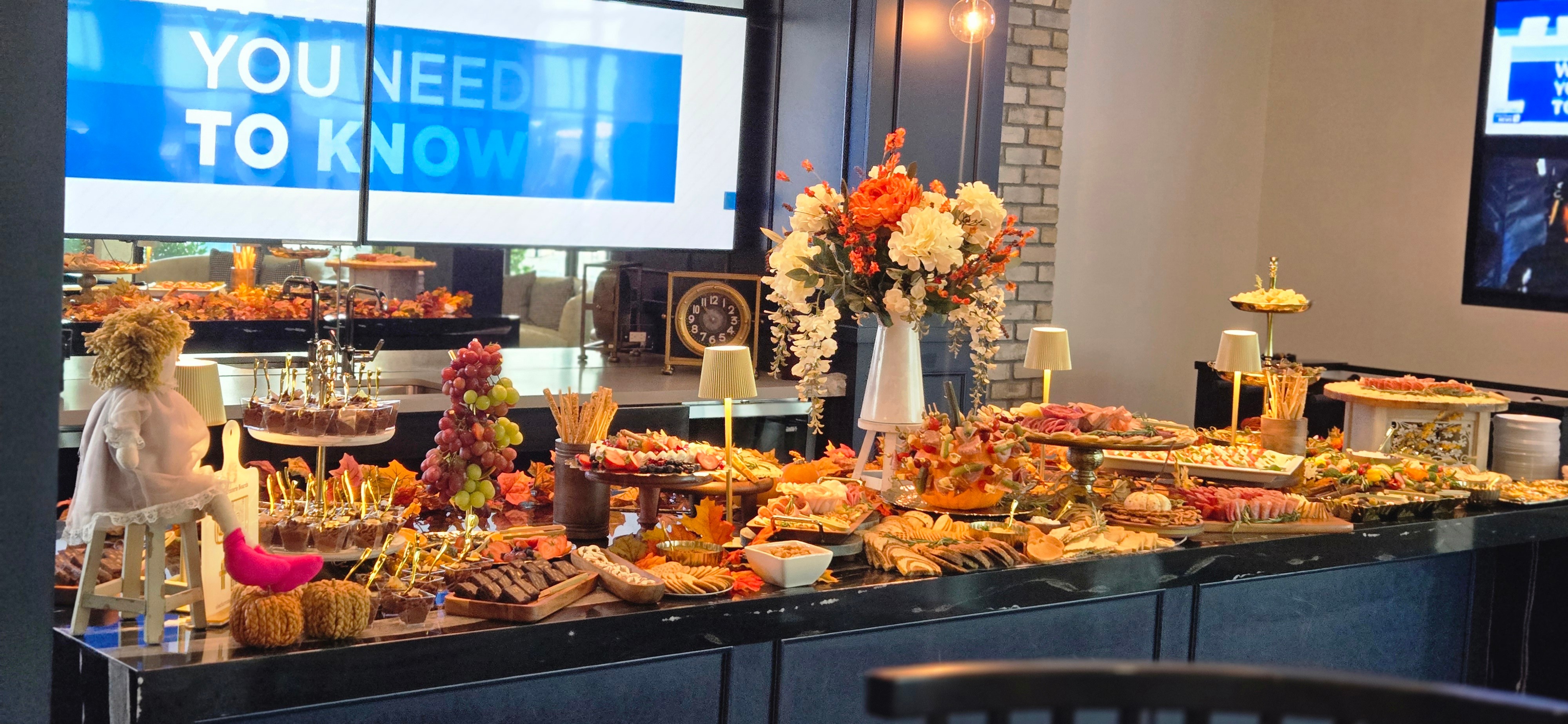 Buffet table with various food, flowers, and decorations in front of a window with a blue sign, in a well-lit room.