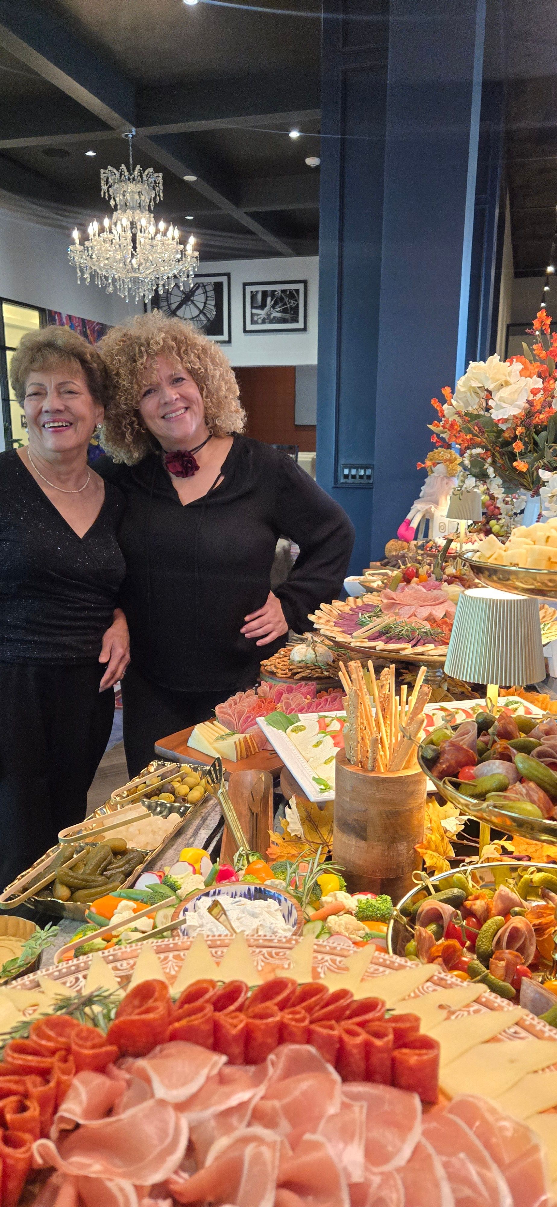 Two women stand beside a decorated table with food and flowers, smiling at the camera in an indoor setting.