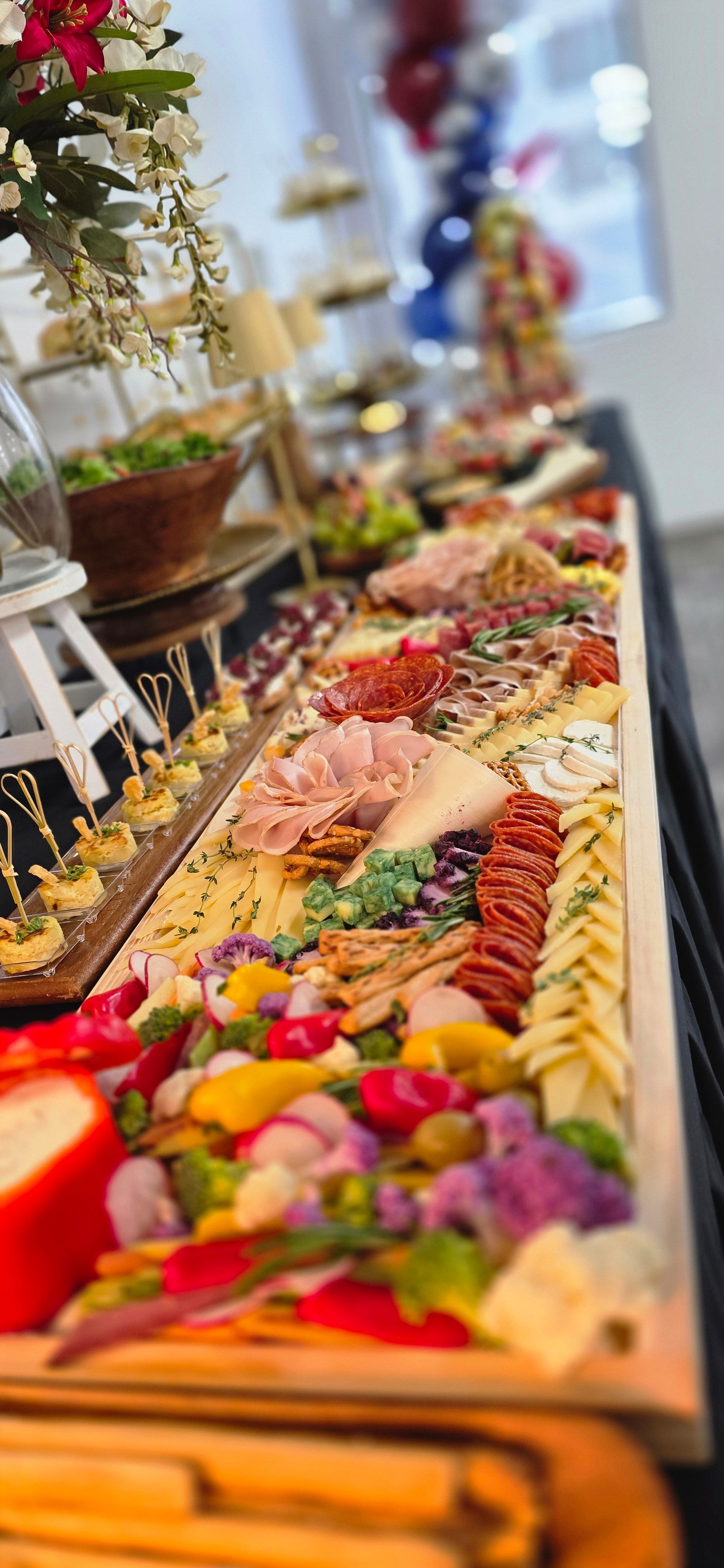 A long table with various colorful desserts and treats, decorated with flowers and greenery, in a bright room.