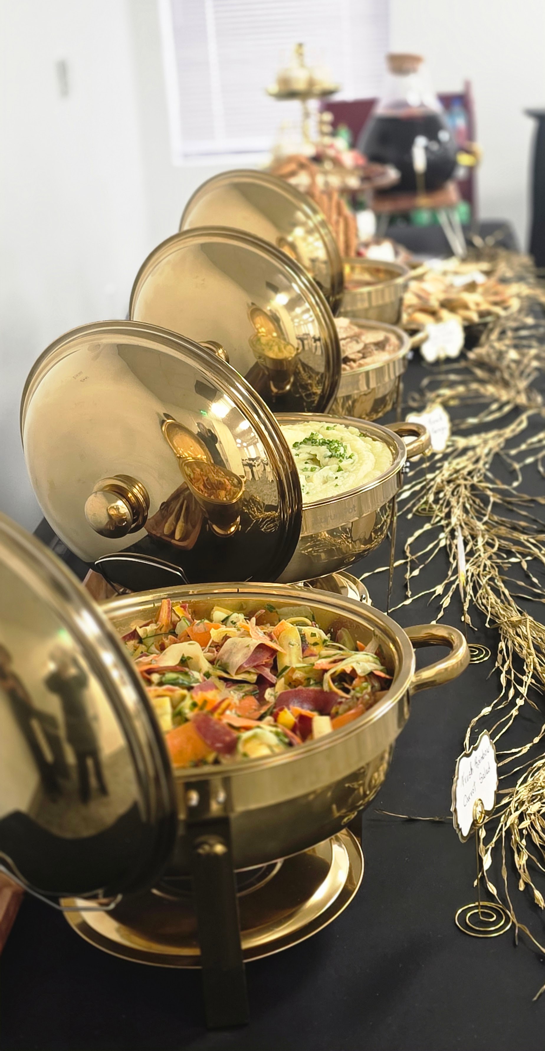 Chafing dishes with food on a black tablecloth, decorated with gold accents and a blurred background.