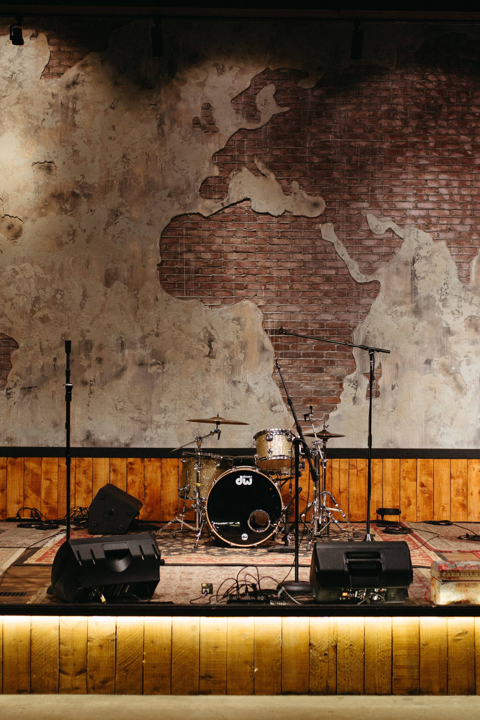 Stage with drum set and microphones in front of a wall featuring a map of South America, illuminated by stage lights.