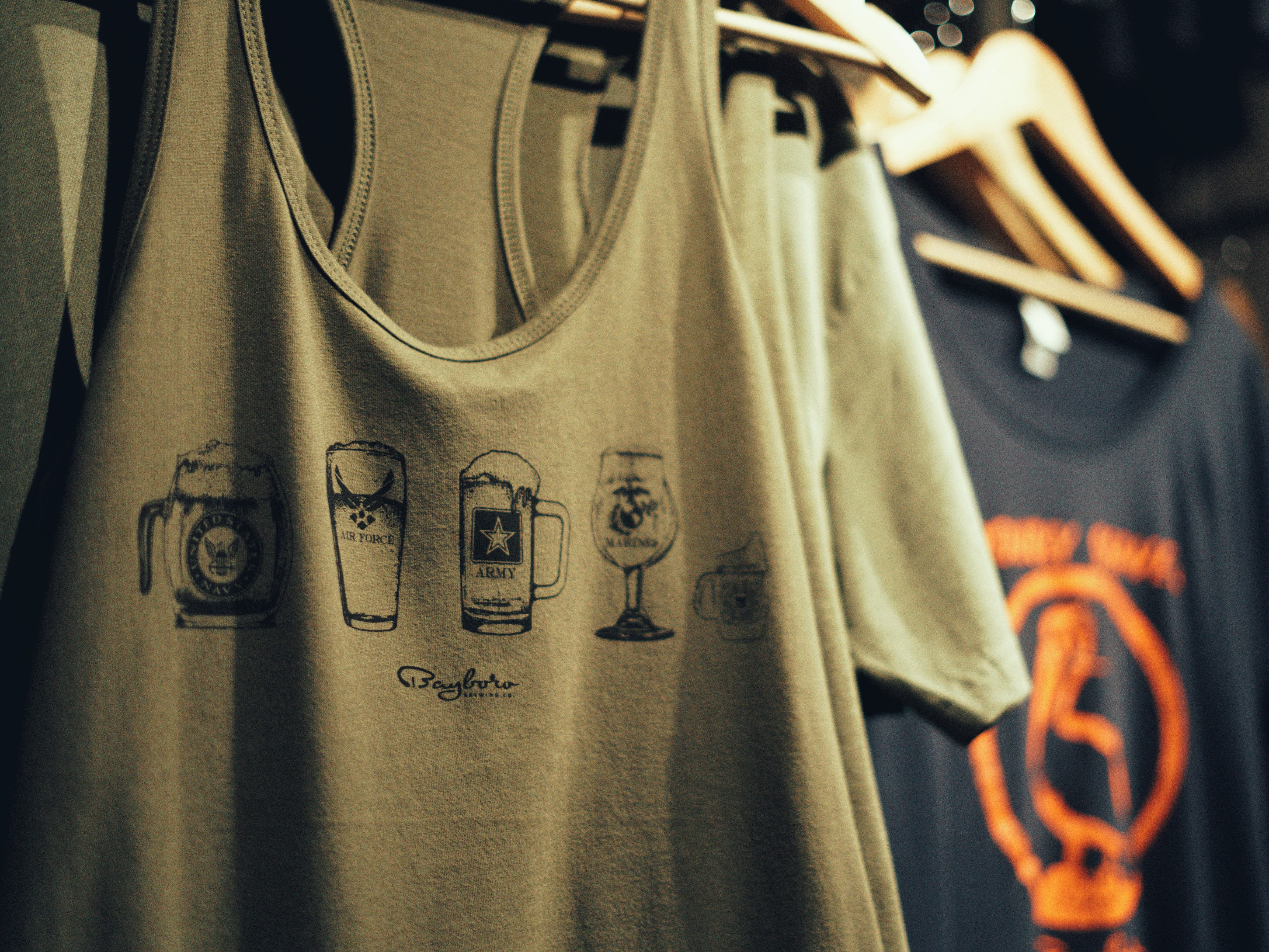 Olive green tank top with beer glass illustrations hanging on a rack, with a black shirt in the background.