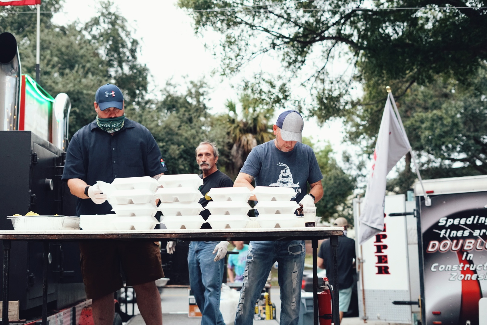 Two men wearing masks and caps stand behind a table with stacked food containers, outdoors with trees and a vehicle in background.