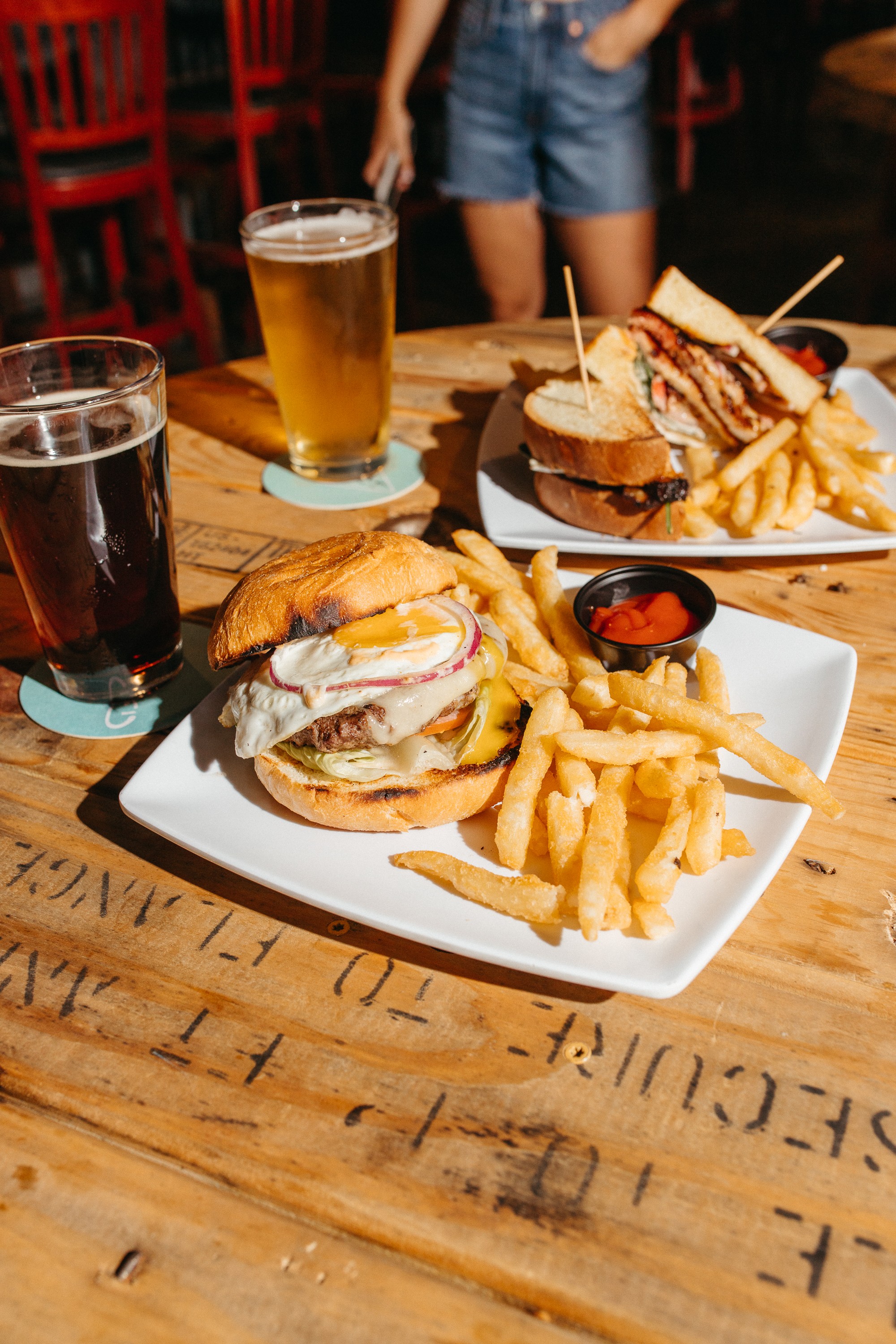 Two plates of burgers with fries, two drinks, and a person in shorts in the background at a wooden table.