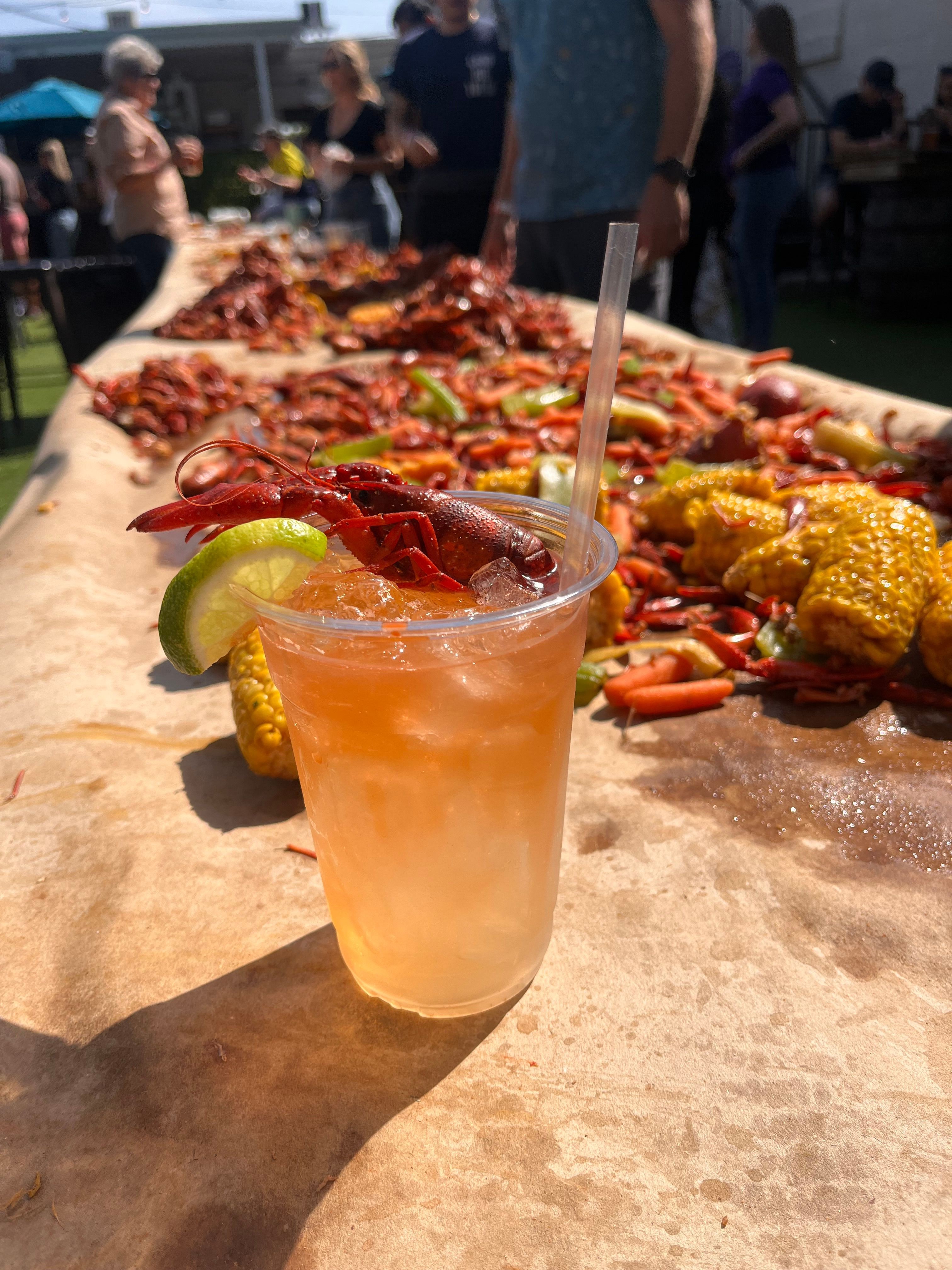A glass of orange-colored beverage with lime and chili slices on a table at an outdoor market.