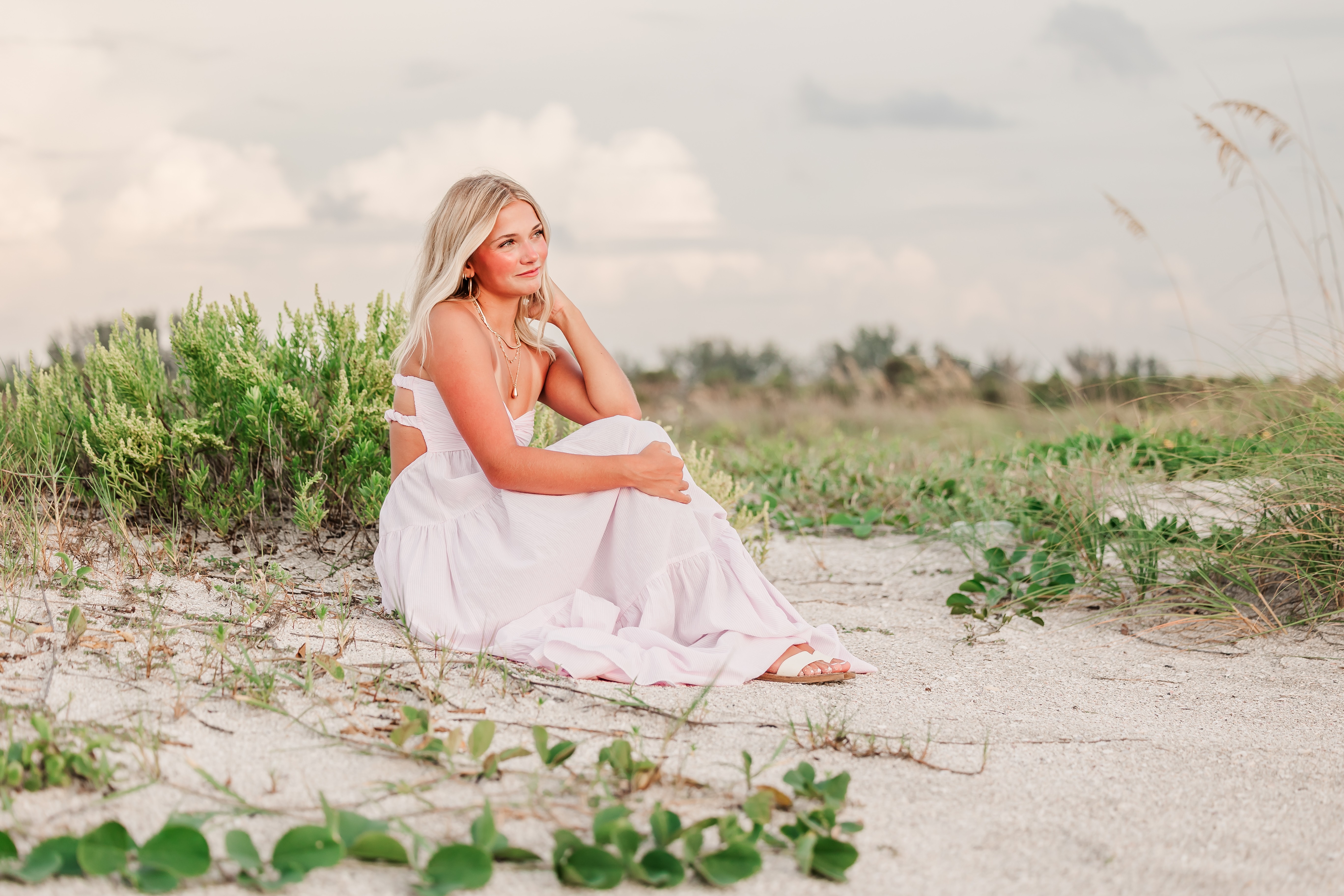 Woman sitting on sandy ground near greenery, looking into distance, wearing a white dress, overcast sky