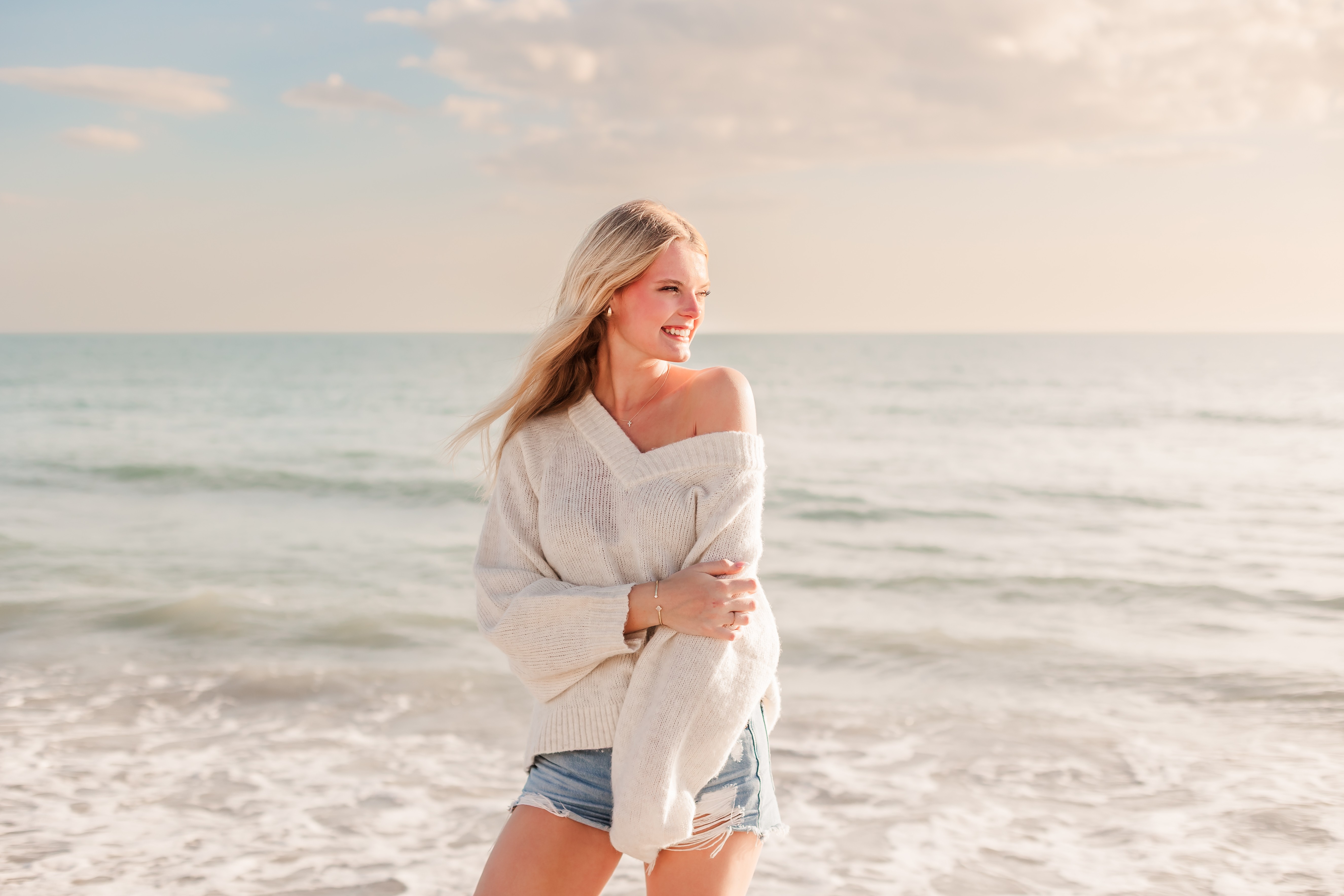 Woman with blonde hair standing on the beach, smiling, wearing a white sweater and denim shorts, ocean in background.