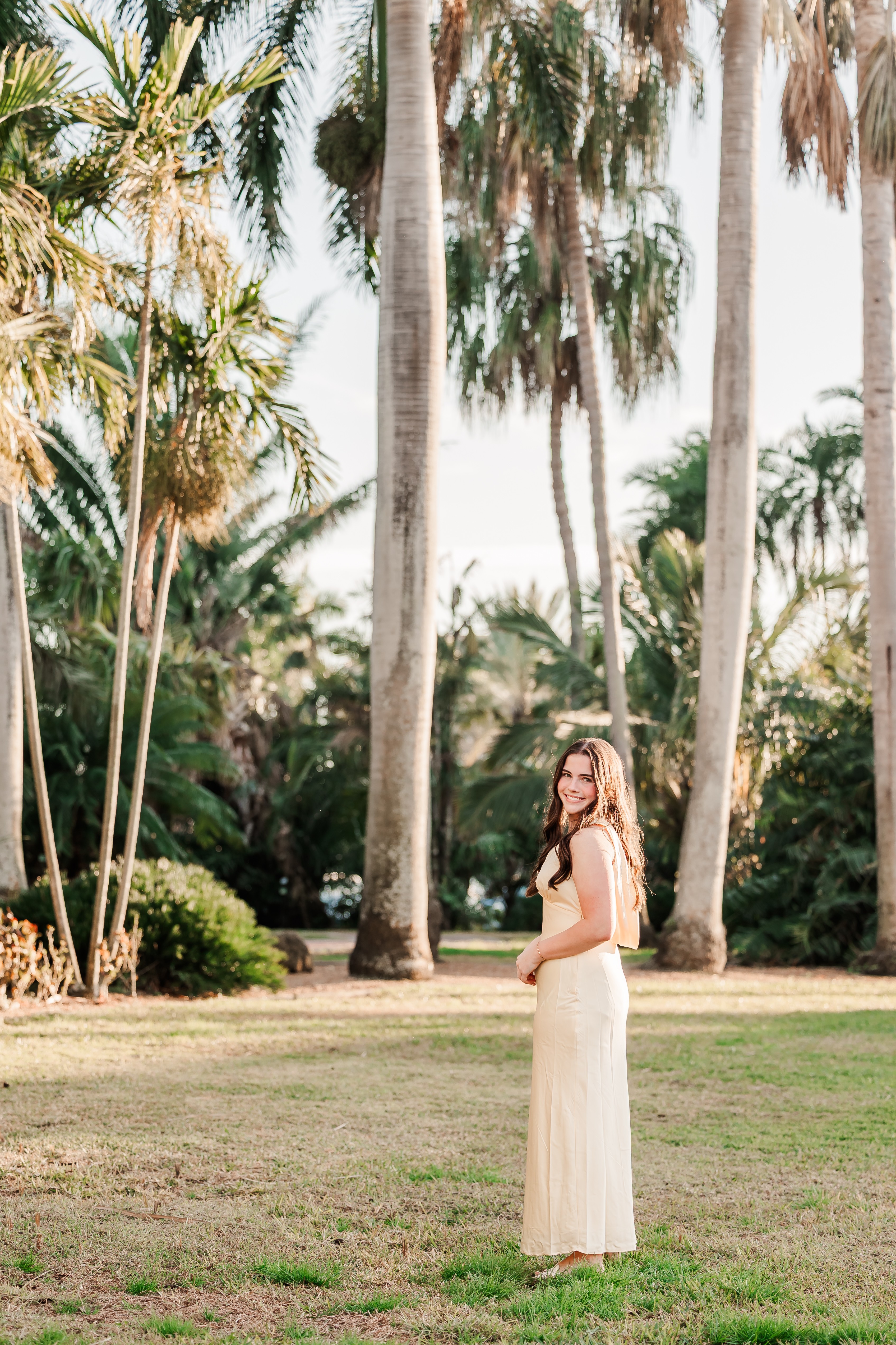 Woman in a long dress standing outdoors among tall palm trees, with greenery and a bright sky in the background.