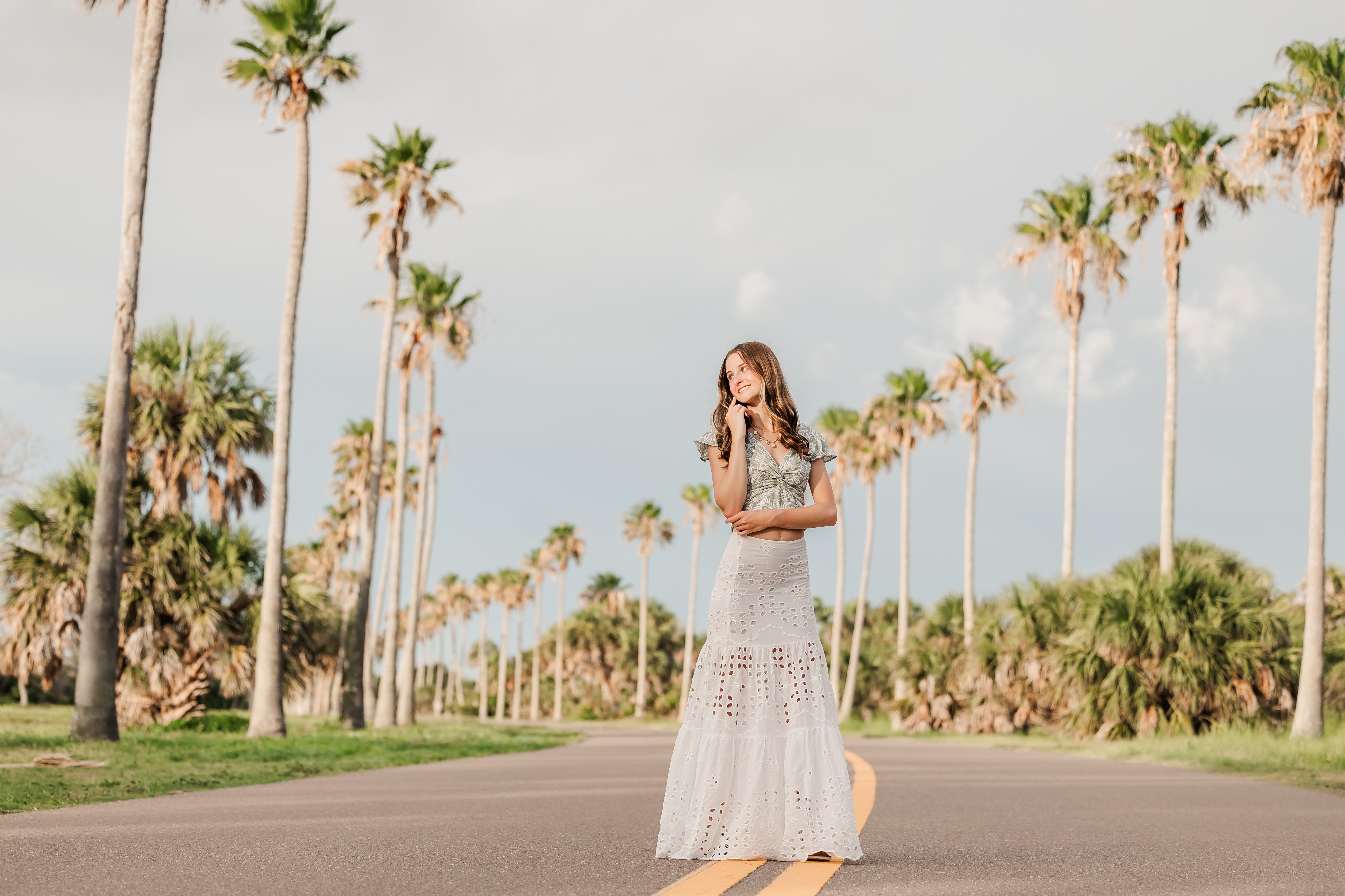 Woman in a long dress standing on a road with palm trees in the background, clear sky, and greenery.