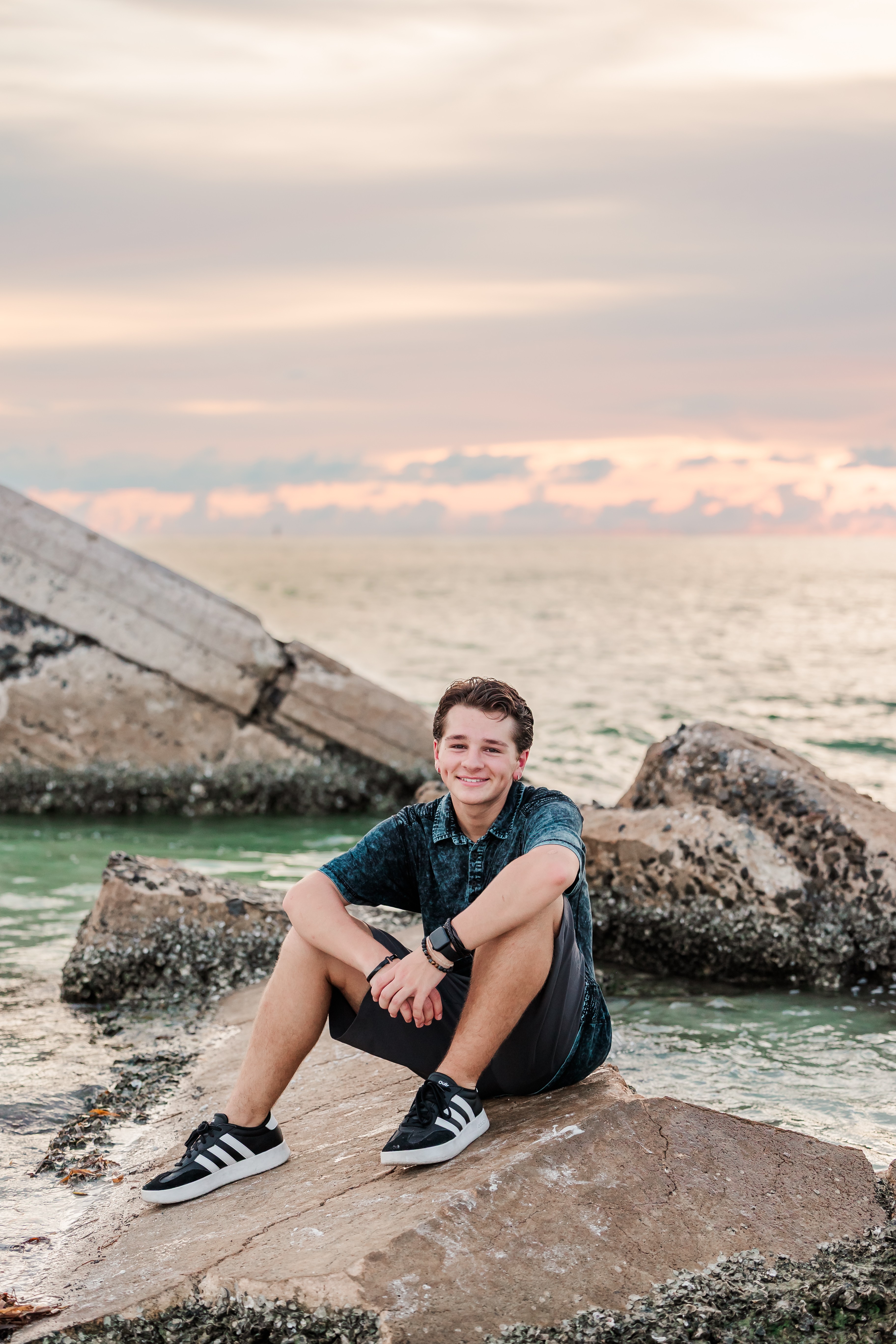 Person sitting on rocks near water during sunset, smiling, wearing a blue shirt and sneakers.