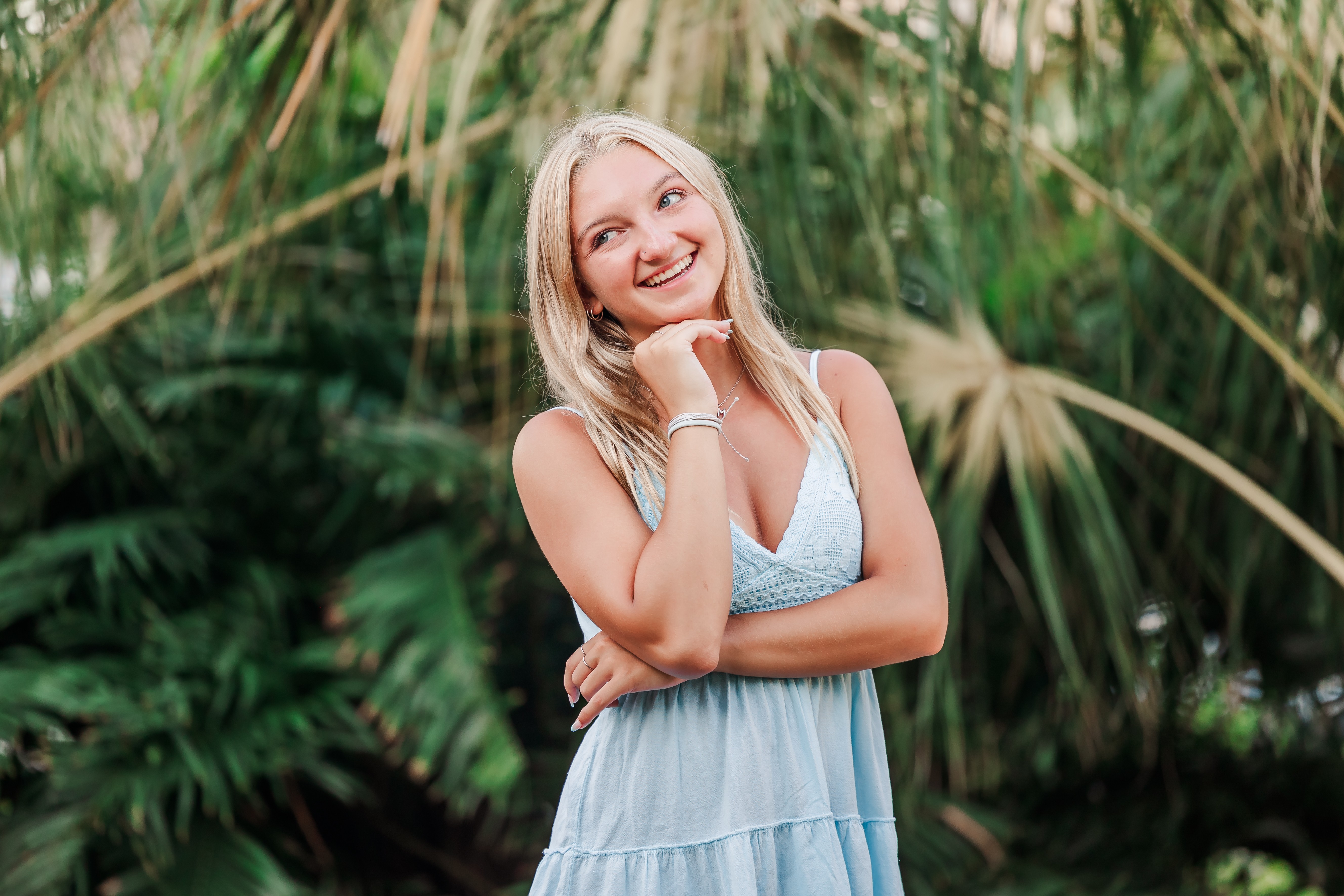 Smiling woman with blonde hair in a light blue dress outdoors with green foliage background.