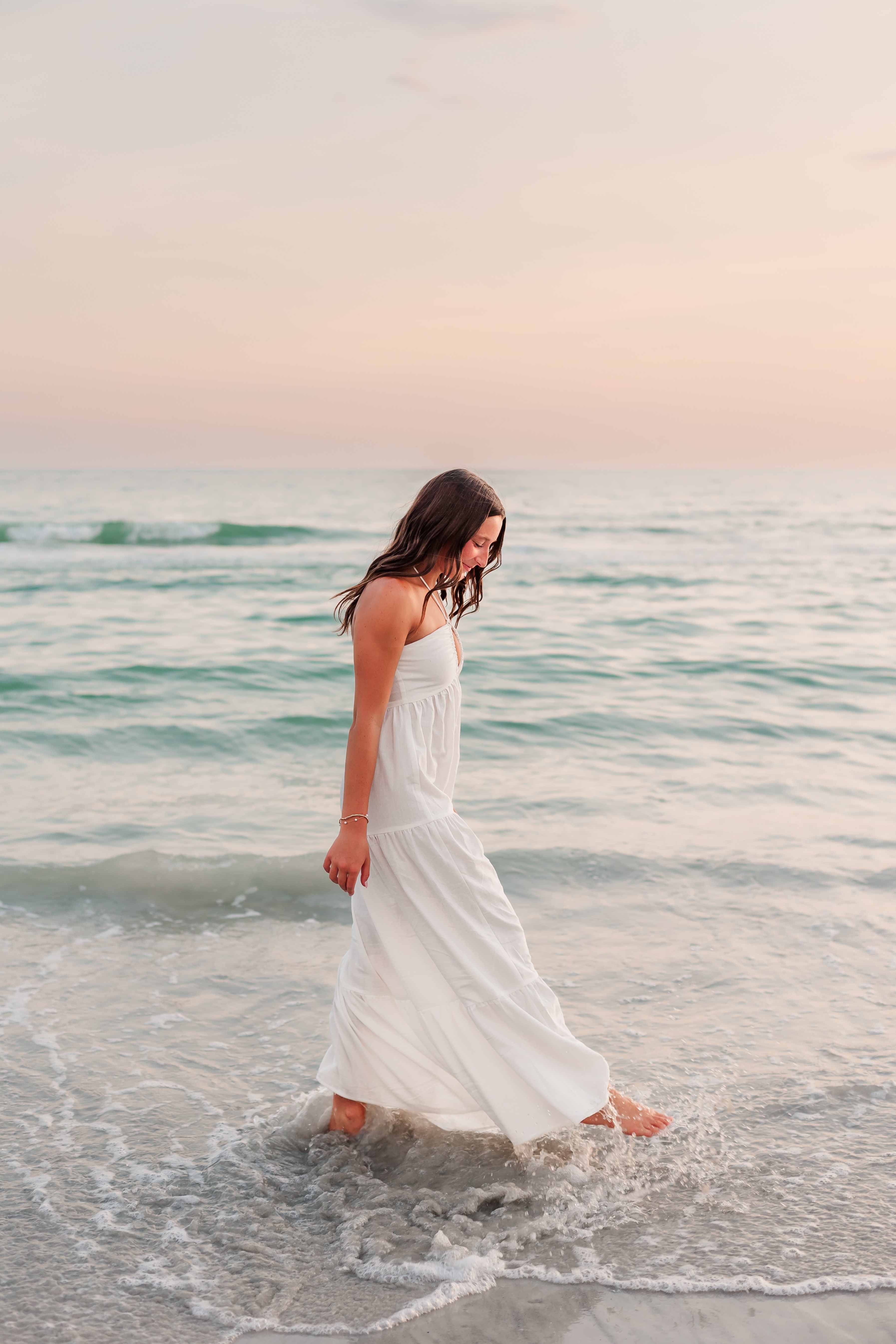 Woman in a white dress walking in shallow ocean water at the beach, with a calm sea and sky in the background.