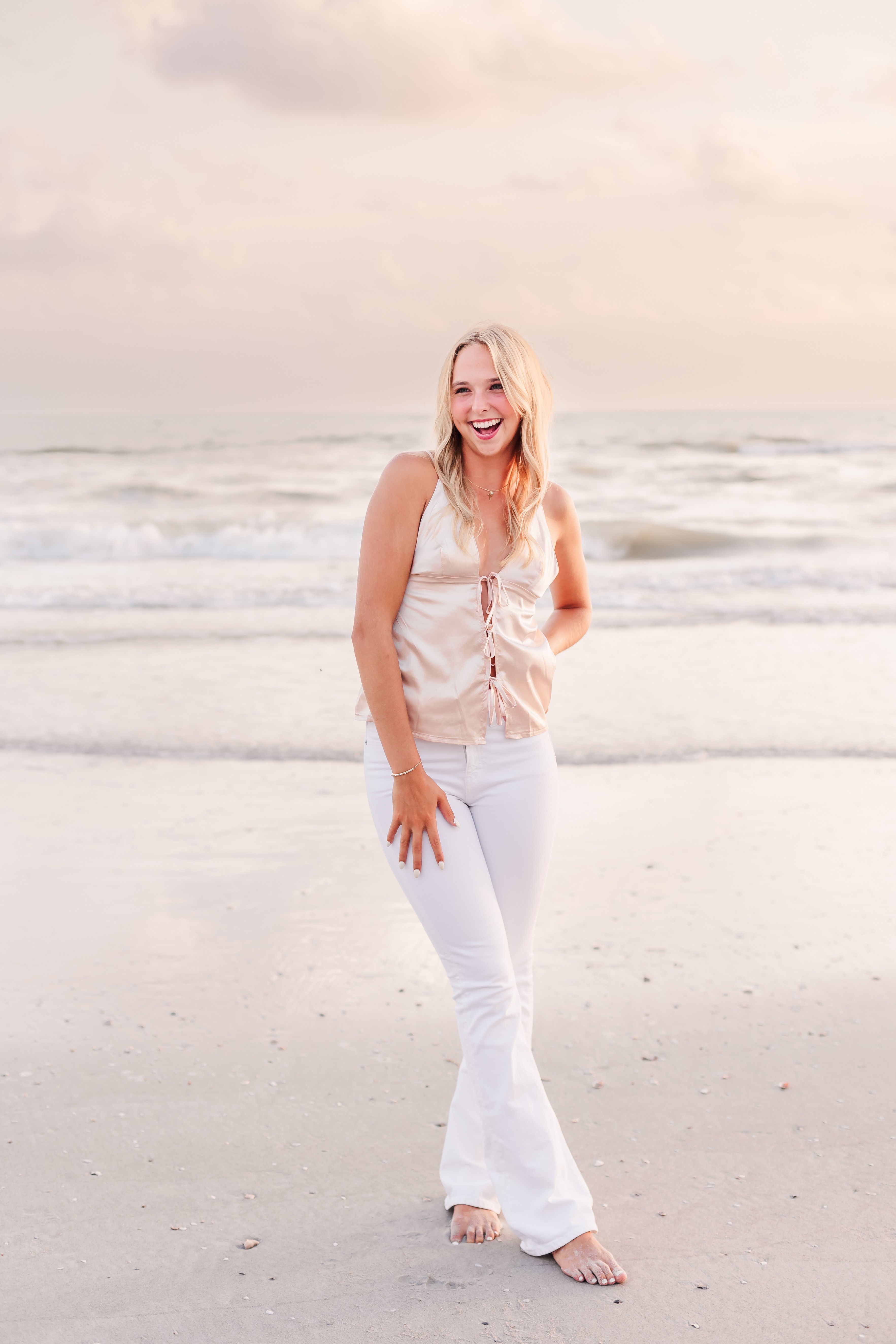 Woman with long blonde hair smiling on beach, wearing white pants and sleeveless top, standing on sand with ocean behind her.