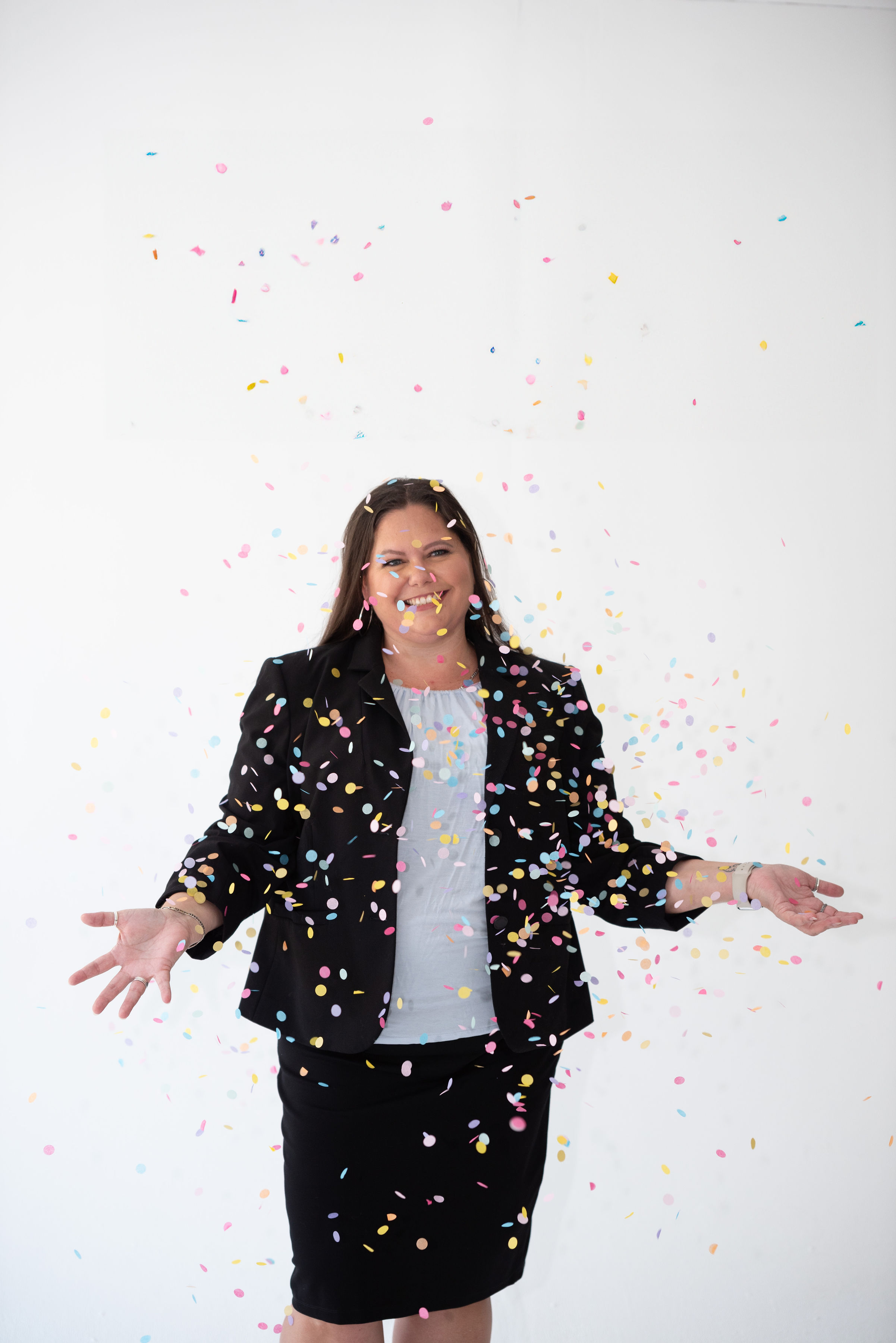 Woman in business attire celebrating with colorful confetti falling around her, smiling, against a plain white background.