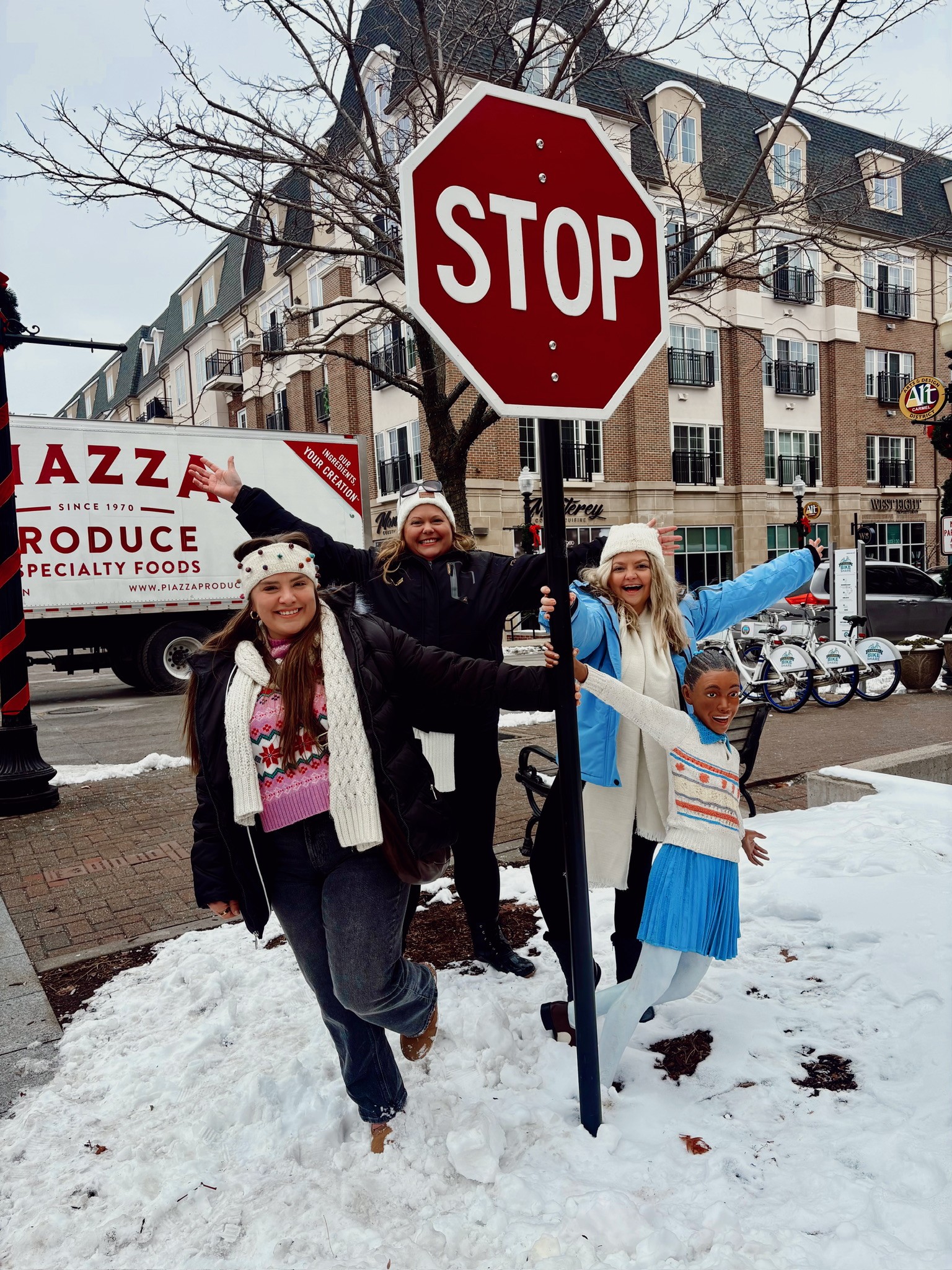 Three children standing near a stop sign on a snowy sidewalk, smiling and pointing, with buildings and bicycles in the background.