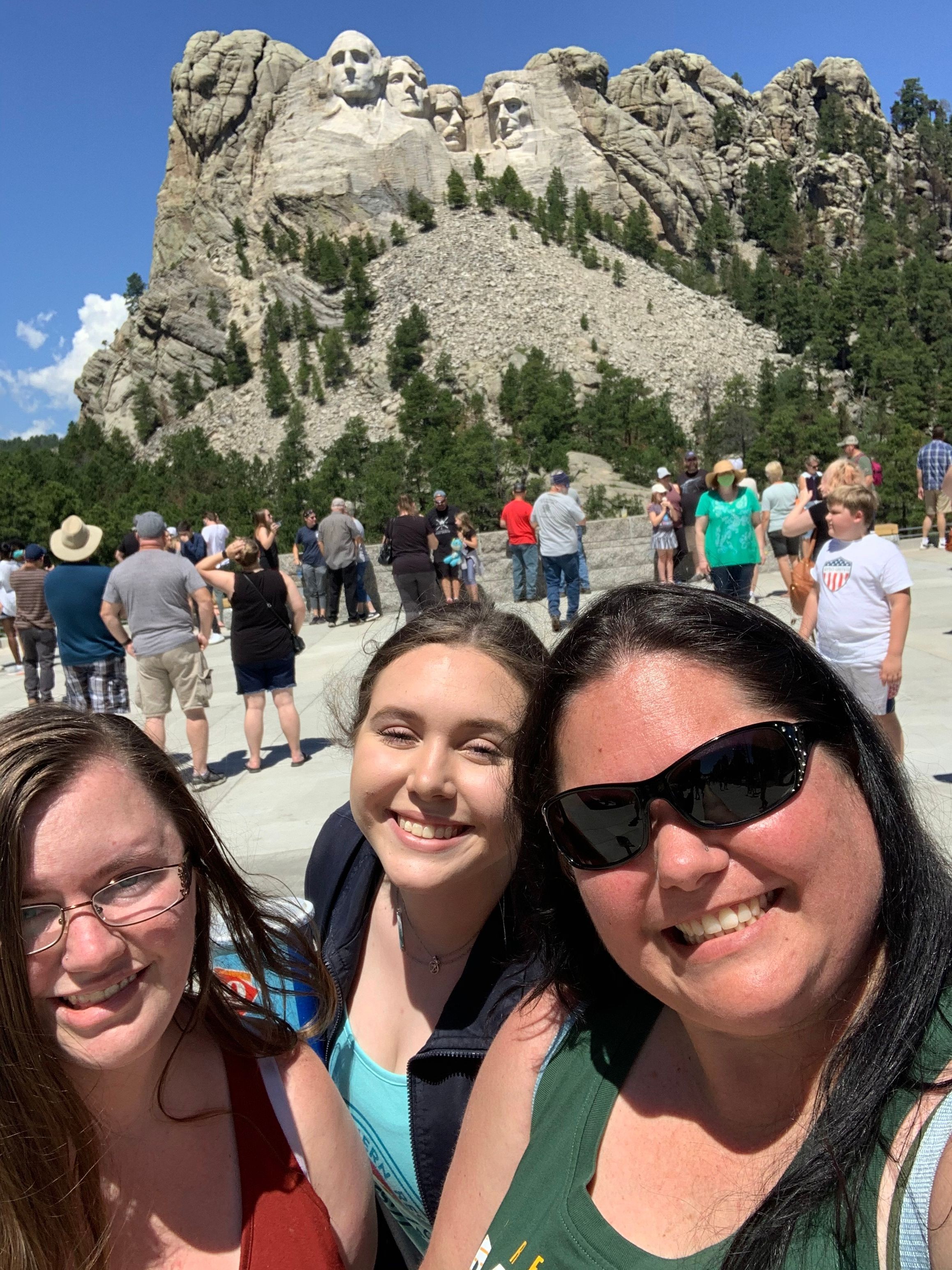 Three women smiling outdoors with a rocky hillside and a crowd of people in the background.