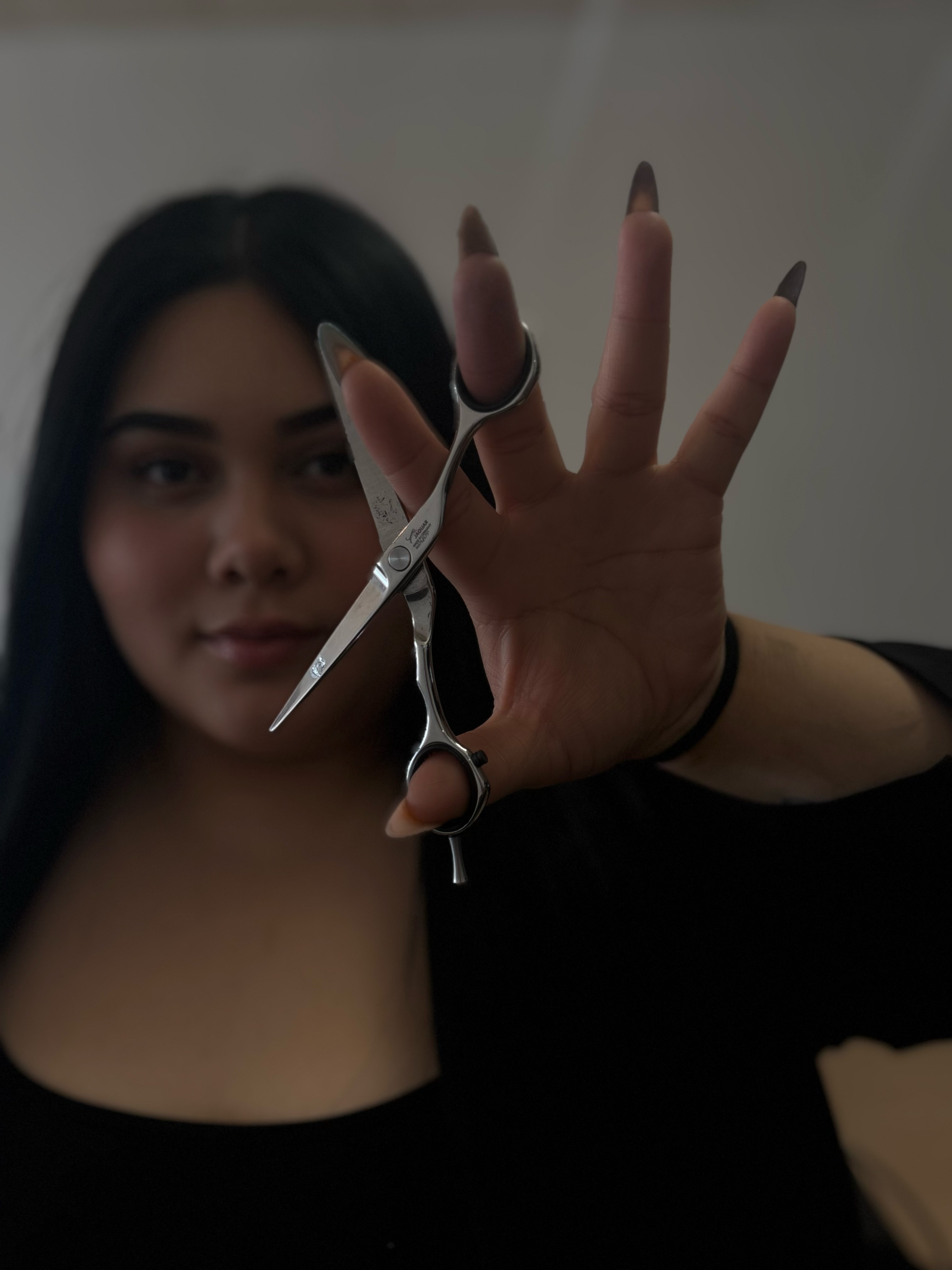 Person with dark hair holding scissors close to camera, hand extended with fingers spread, background blurred.