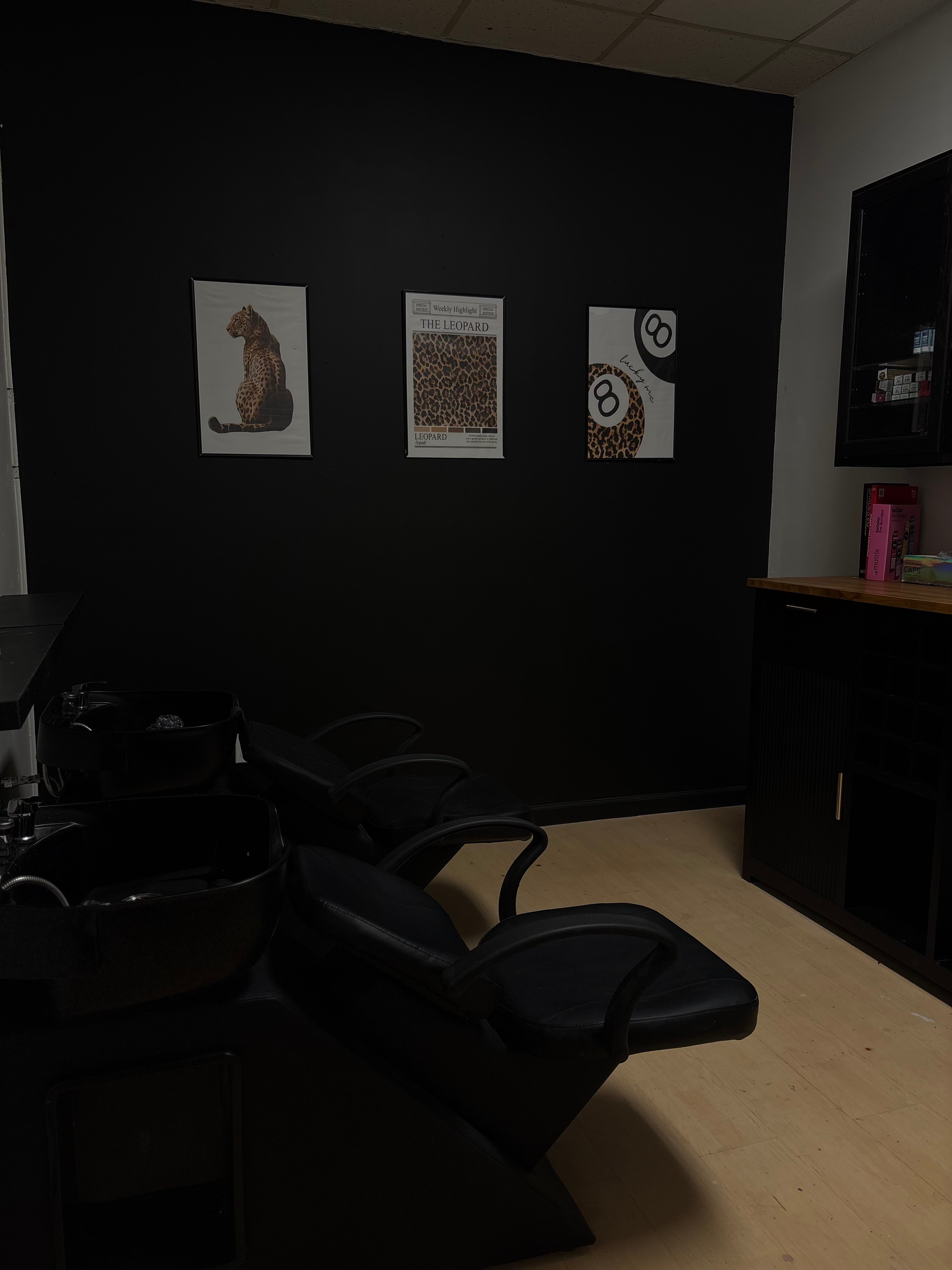 Black wall with three framed pictures, a black chair, and a cabinet with pink and black items.