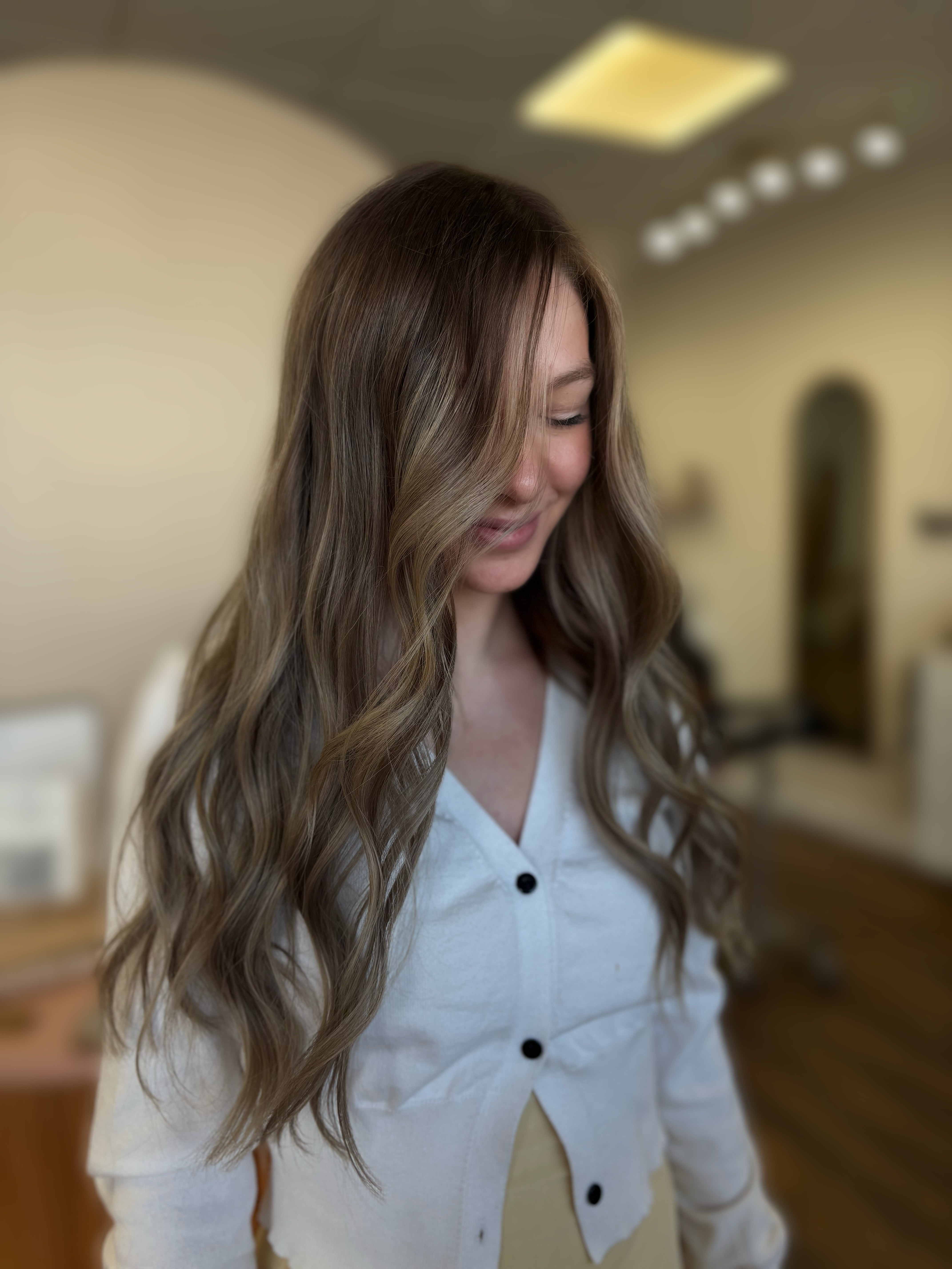 Young woman with long wavy hair looking down, wearing a white shirt, in a room with warm lighting.
