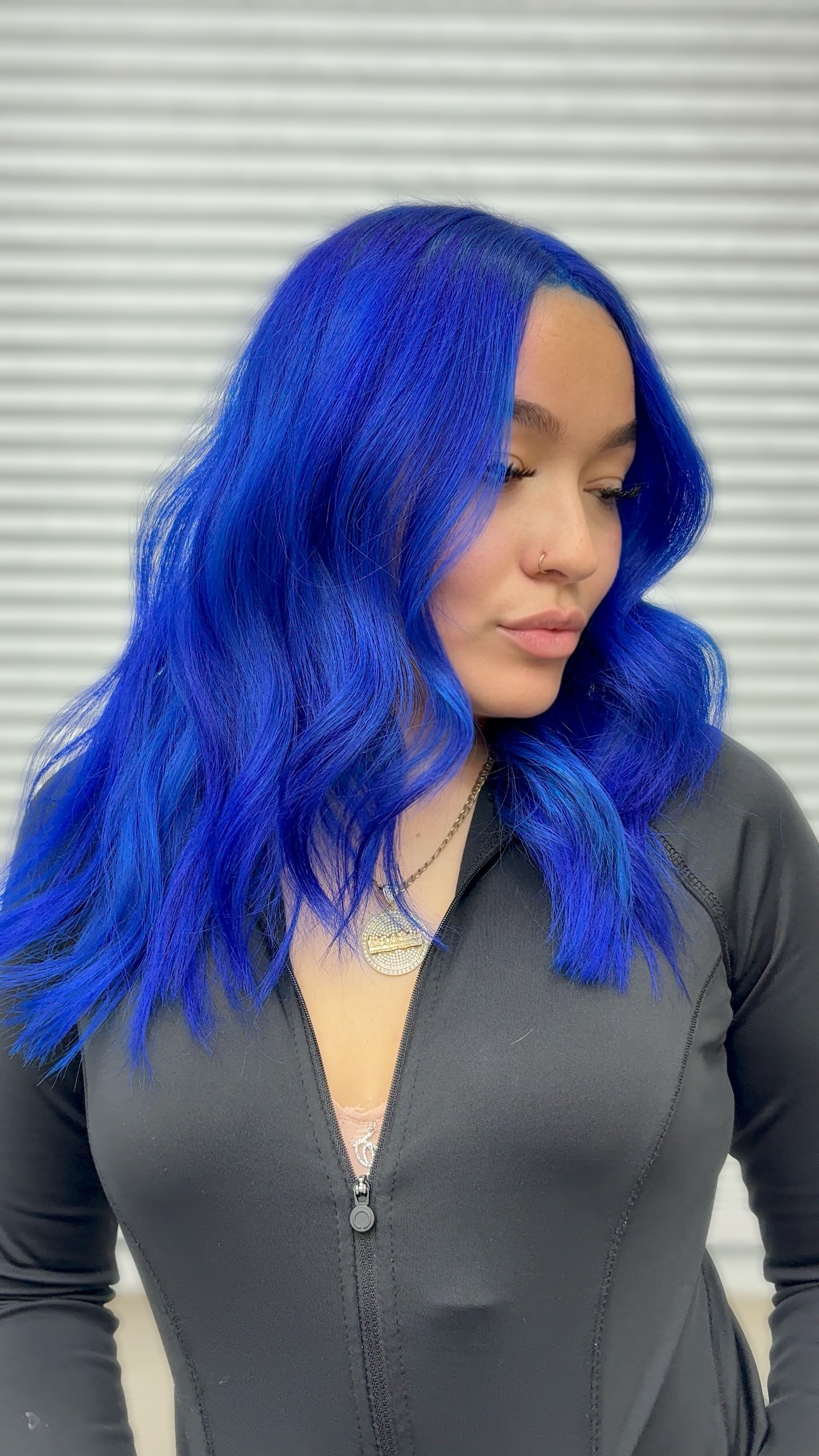 Woman with long, wavy, bright blue hair looking downward, wearing a dark gray zip-up top, background with horizontal lines.