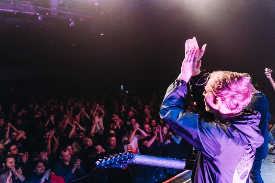 Performer on stage with hands together, facing a large audience in a dark venue.