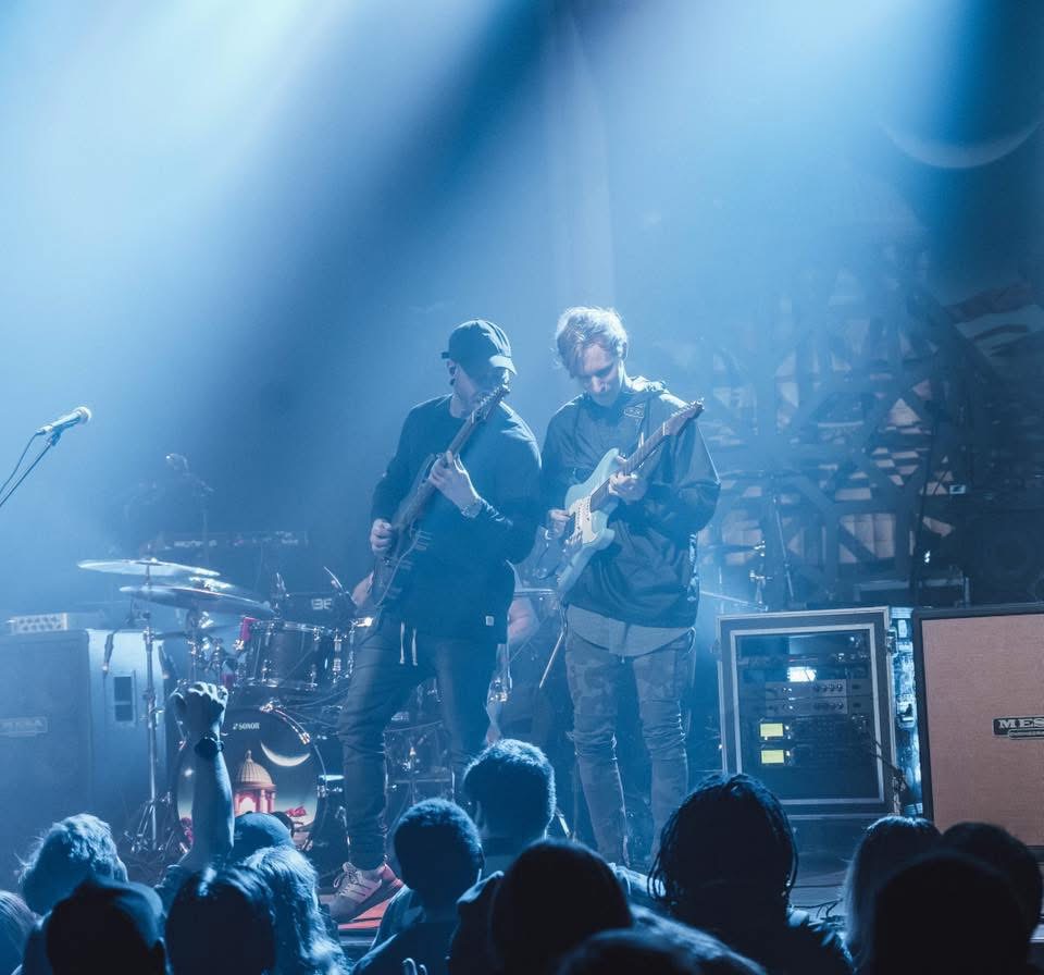 Two musicians perform on stage with guitars, audience visible in foreground, blue lighting, equipment in background.