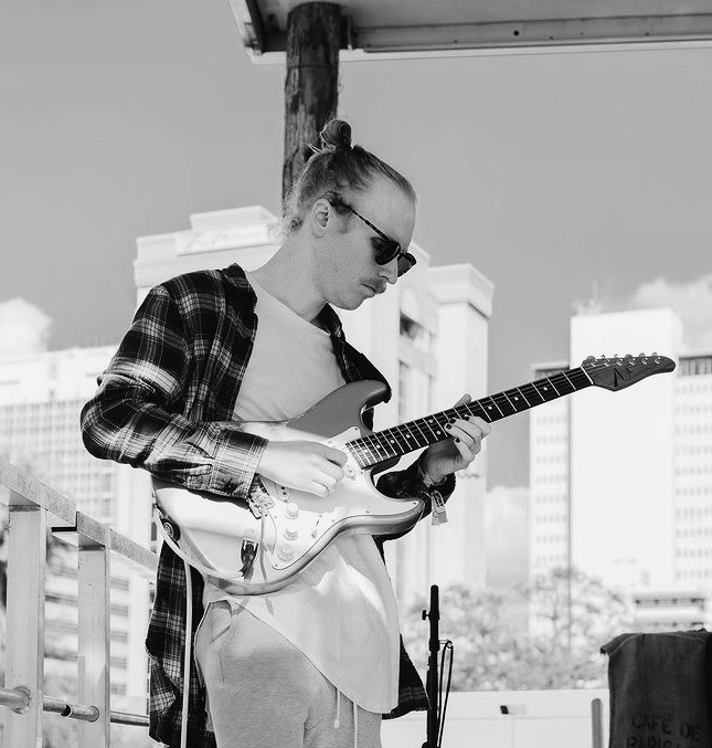 Person playing electric guitar outdoors, wearing sunglasses and a plaid shirt, with city buildings in background.