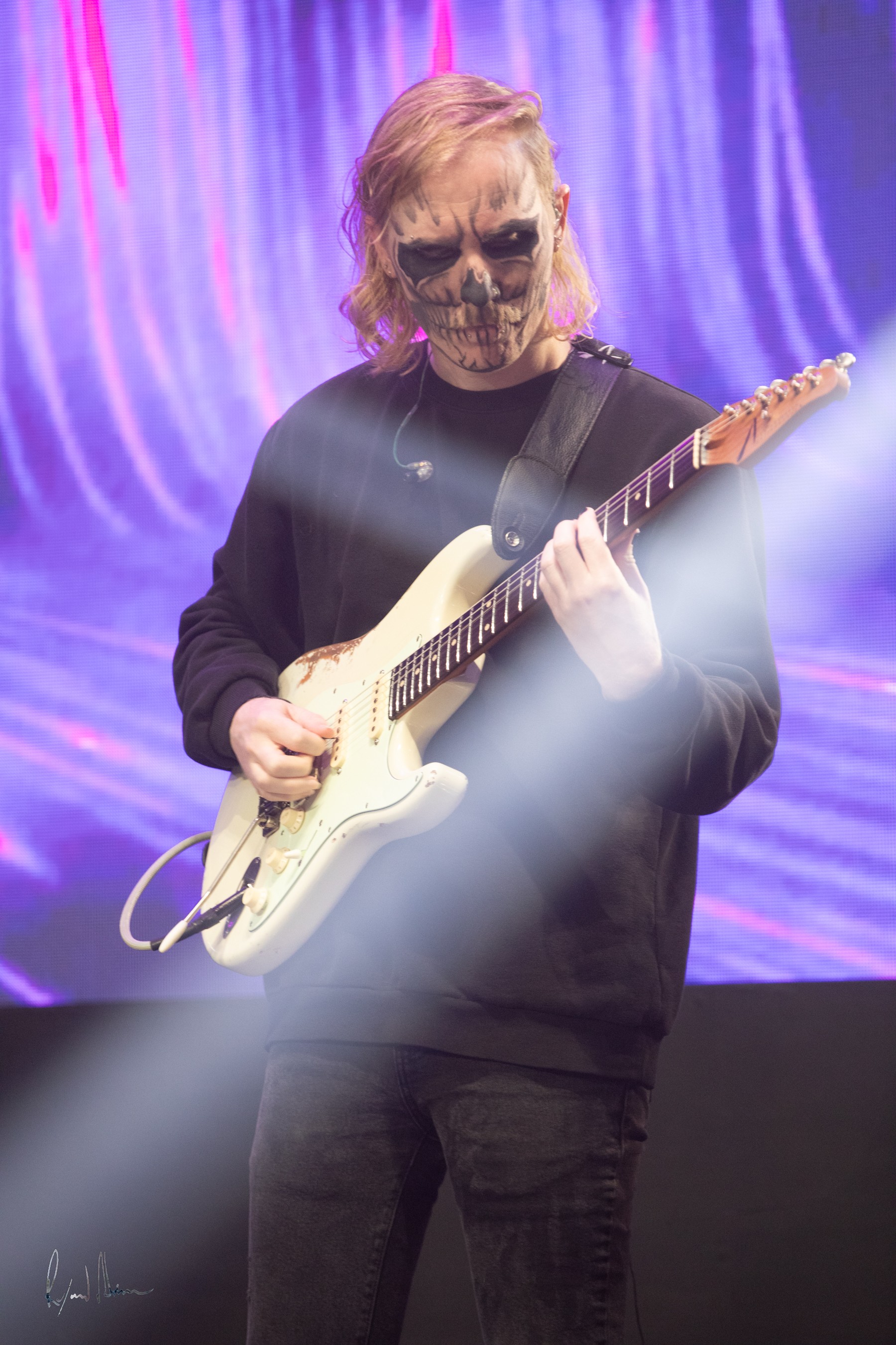 Person with skull makeup playing an electric guitar on stage with purple and blue lighting