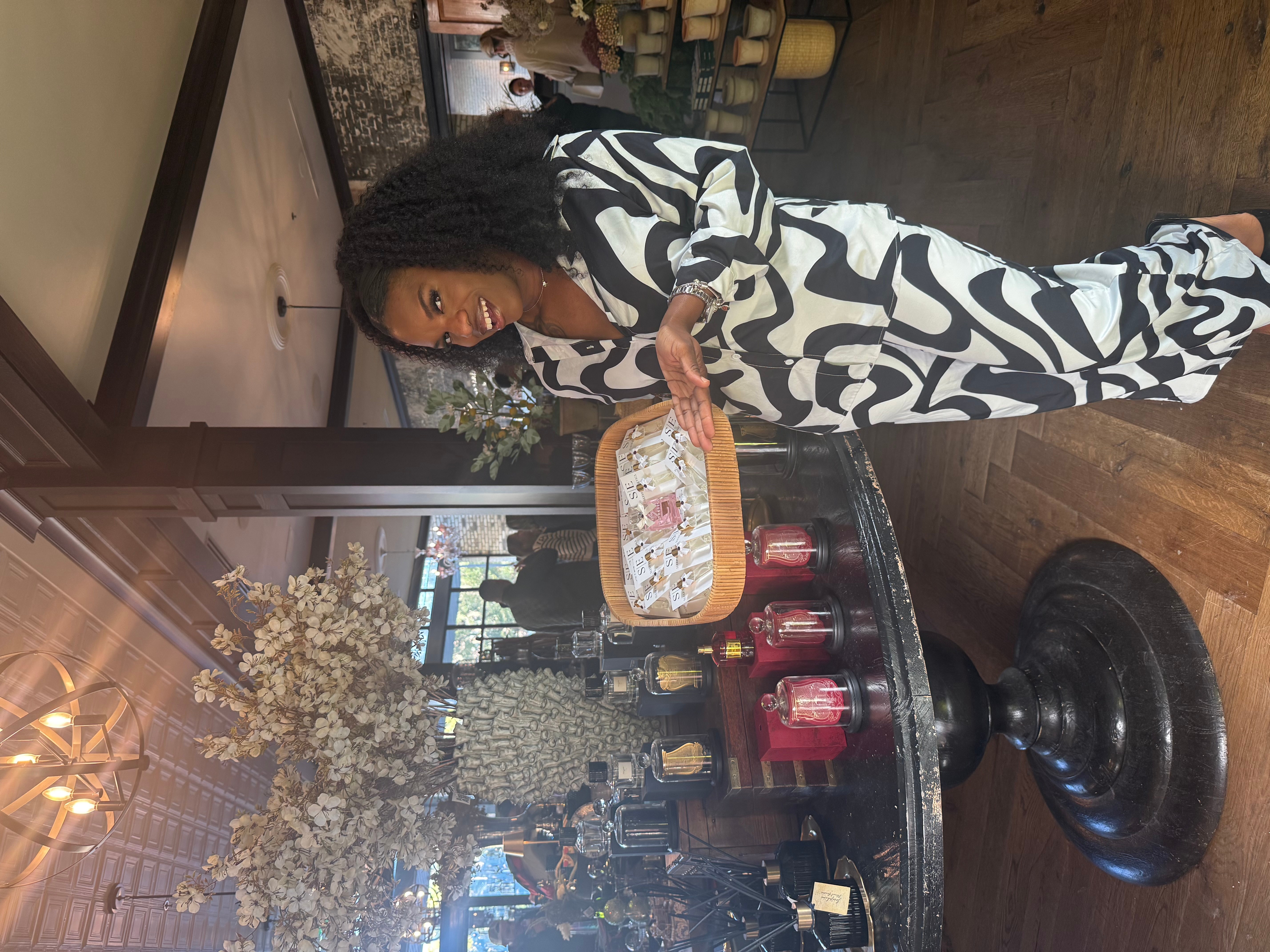 Woman in black and white patterned dress holding a tray with food, standing near a table with candles and flowers.