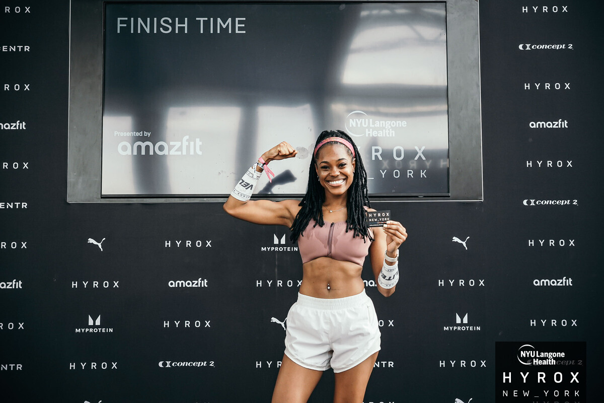 Smiling woman in sportswear flexes arm in front of a large screen with 'Finish Time' text, black background with brand logos.