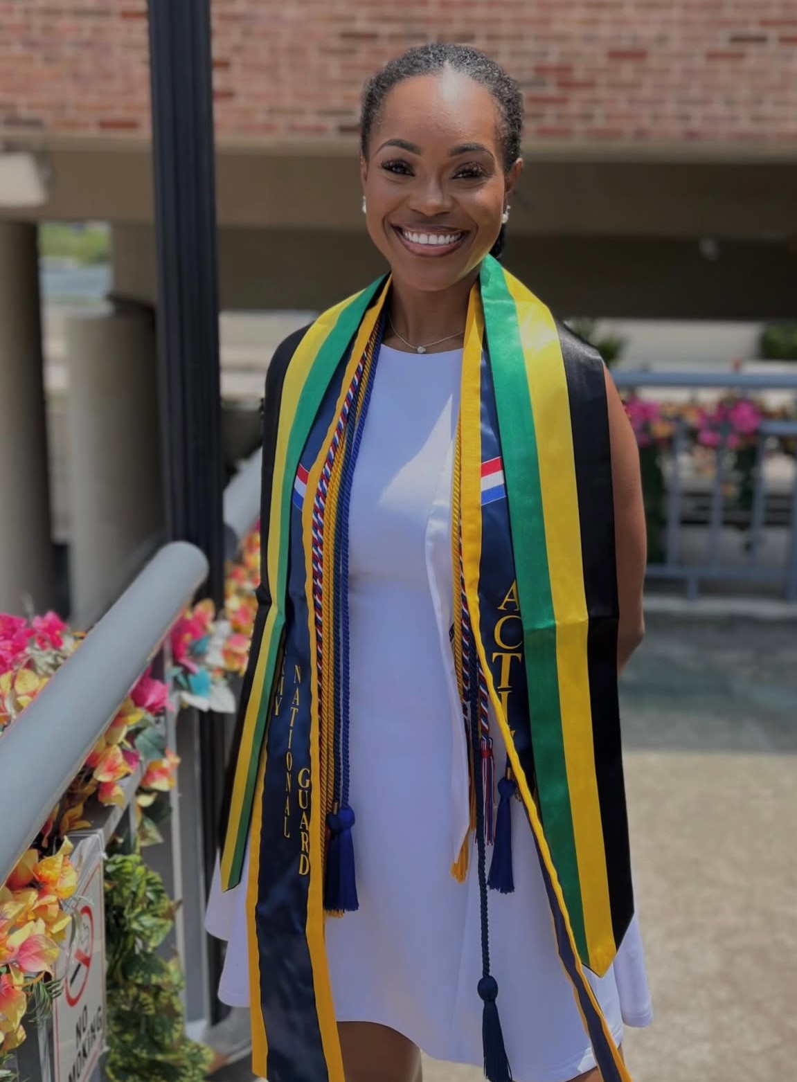 Smiling woman in white dress with graduation stole and cords, standing outdoors near flowers and railing.