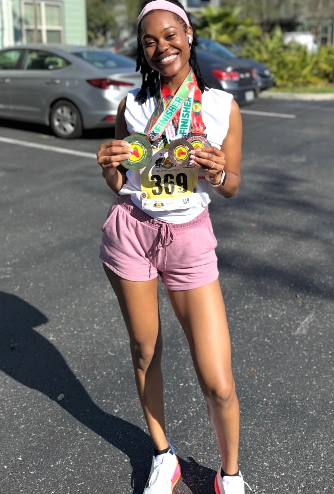 Smiling woman holding medals, wearing a race bib, pink shorts, and running shoes, standing outdoors in a parking lot.