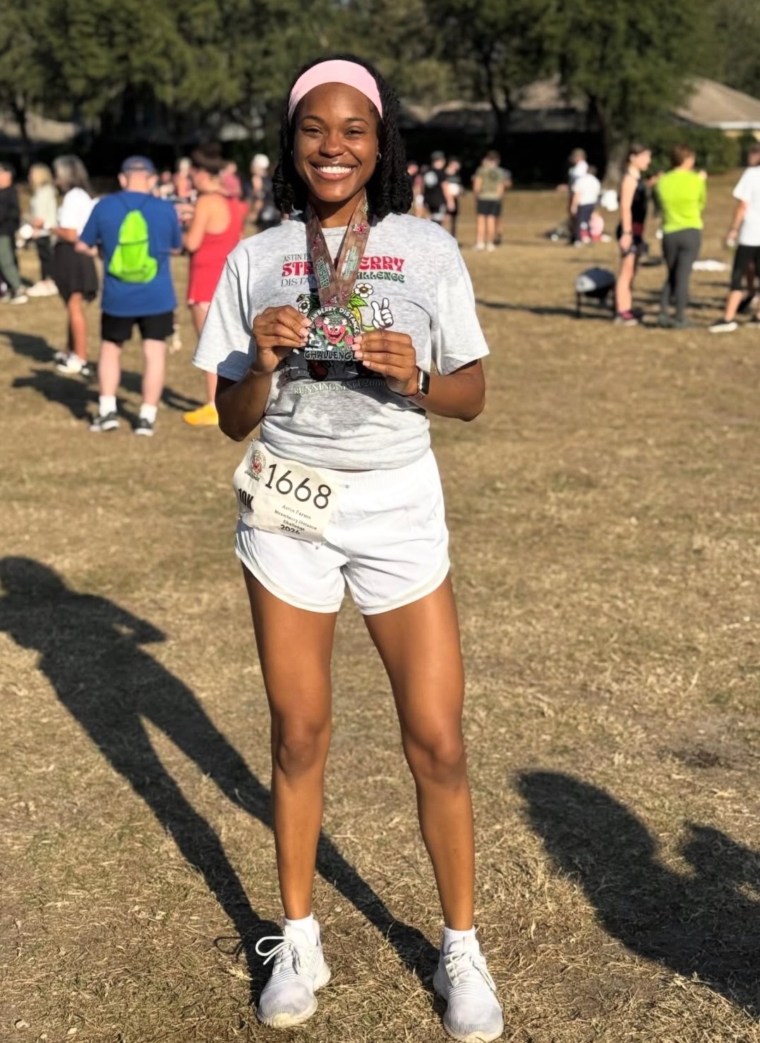 Young woman smiling, holding a medal, standing outdoors with people in the background, wearing athletic clothing and a pink headband.