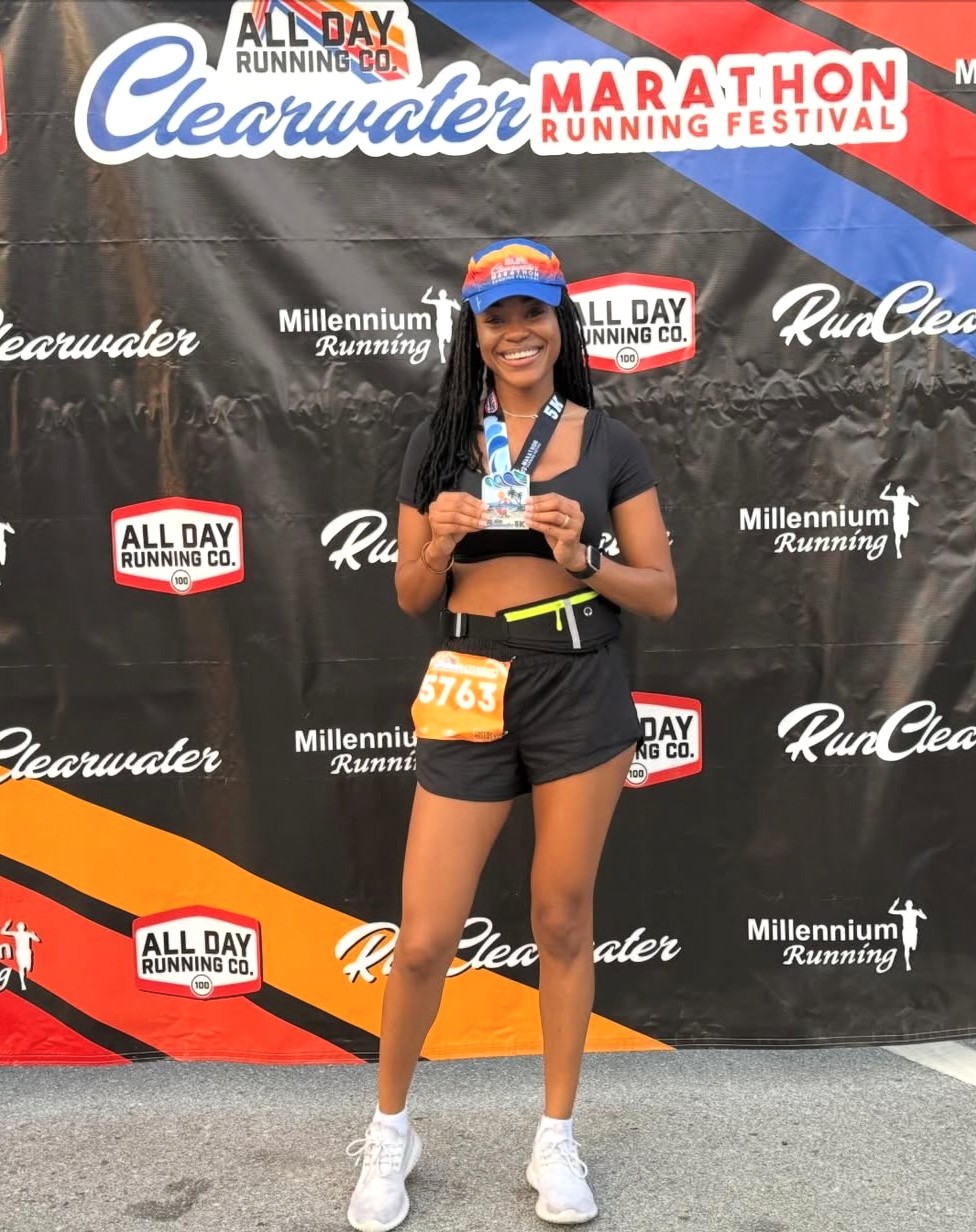 Young woman holding a medal and smiling at a marathon event, standing in front of a black backdrop with event logos.
