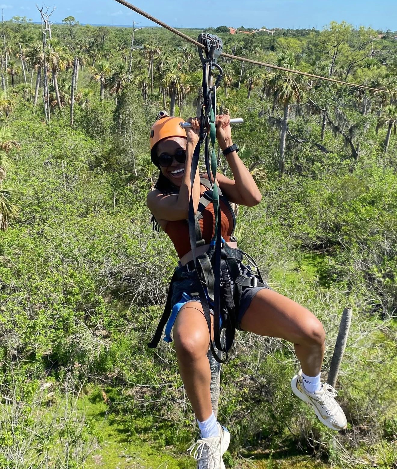 Person wearing helmet and harness, ziplining over green landscape with trees and blue sky, smiling and holding the zipline handle.