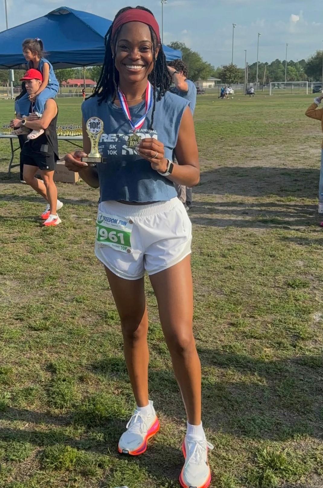 Smiling woman holding a medal and a water bottle at an outdoor event, wearing athletic clothing and a headband.