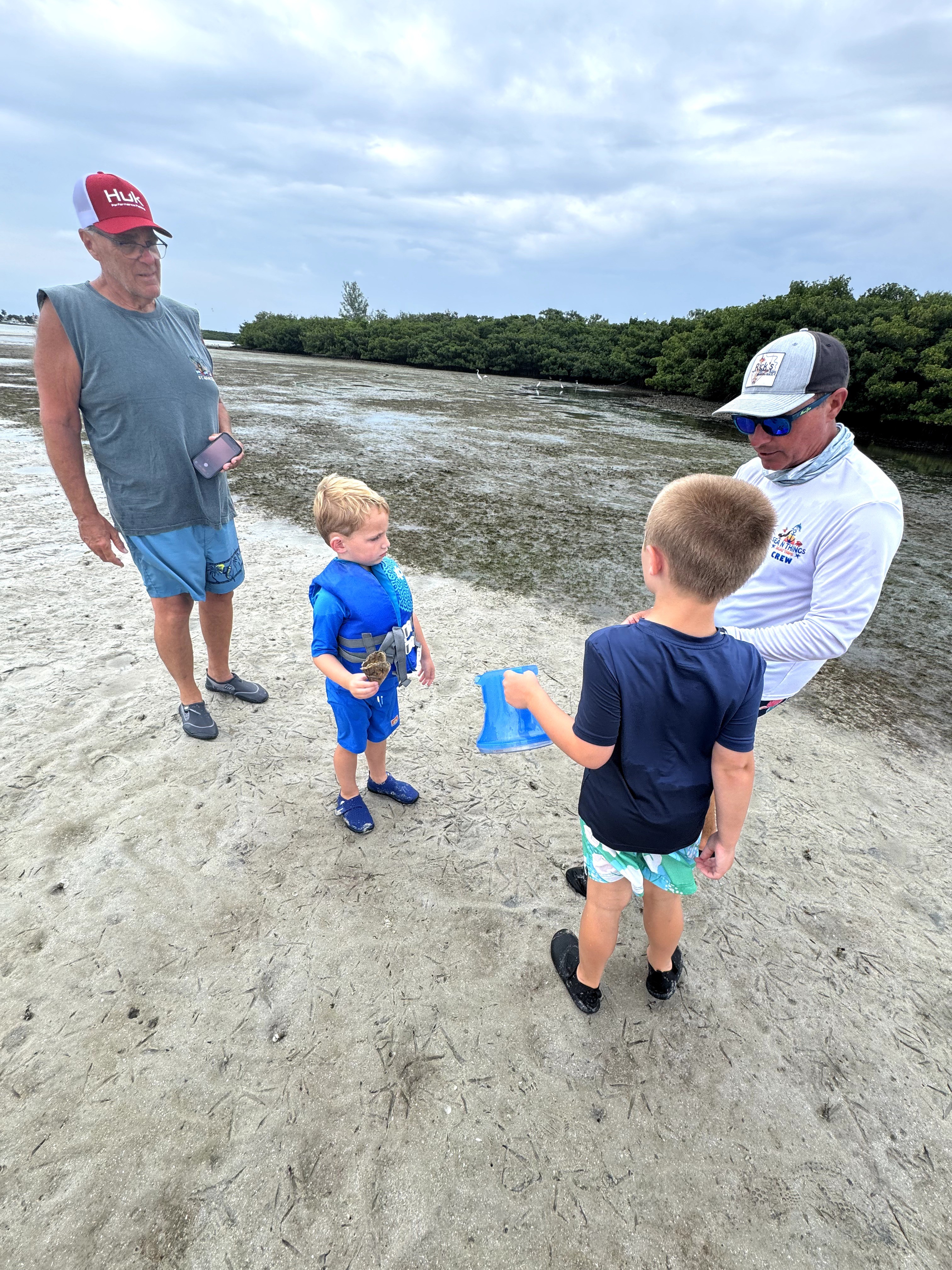 Two children and two adults on a sandy beach near water, with trees and cloudy sky in background.