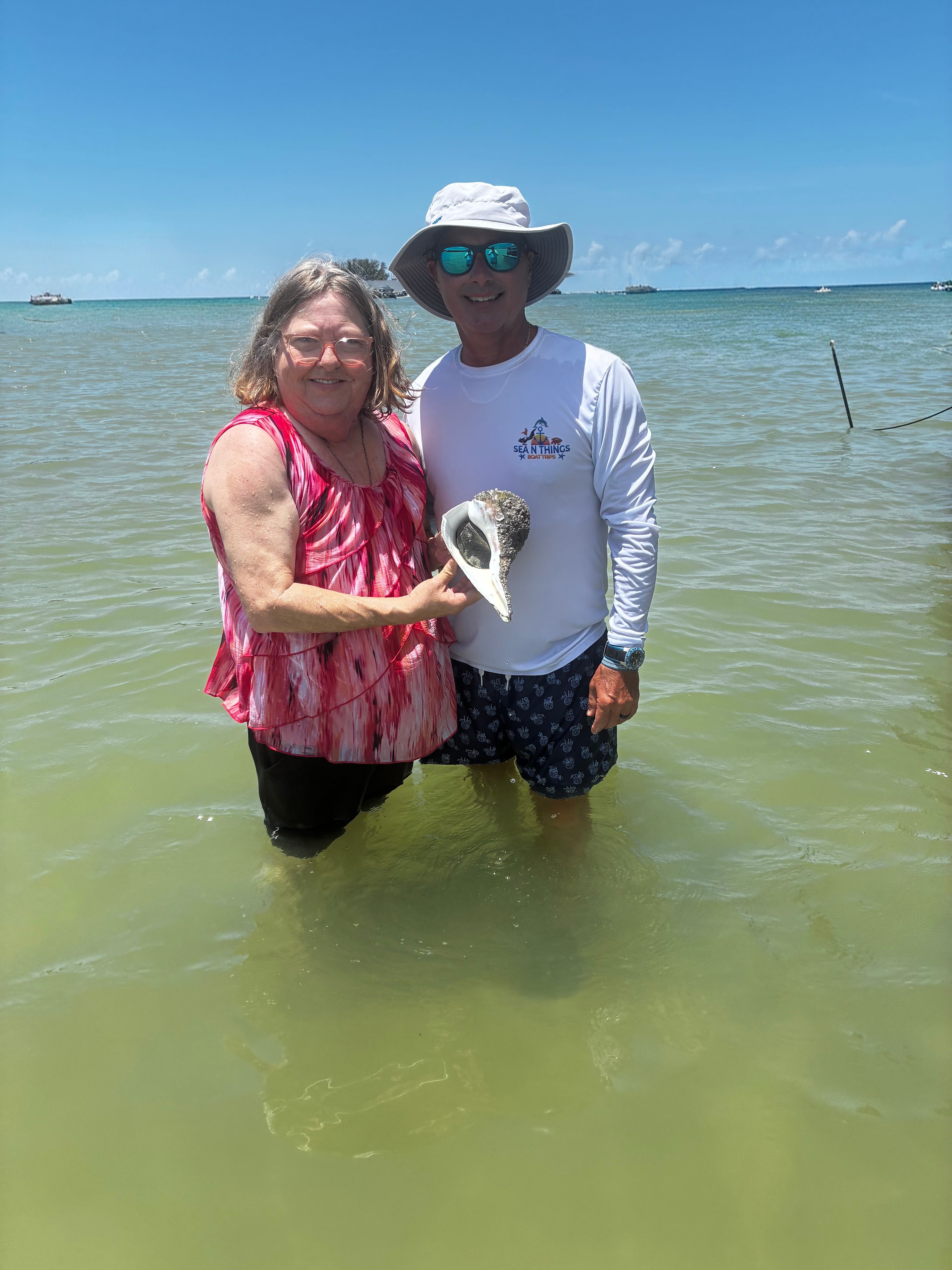 Two people standing in shallow water at the beach, holding a seashell, with a blue sky and ocean in the background.