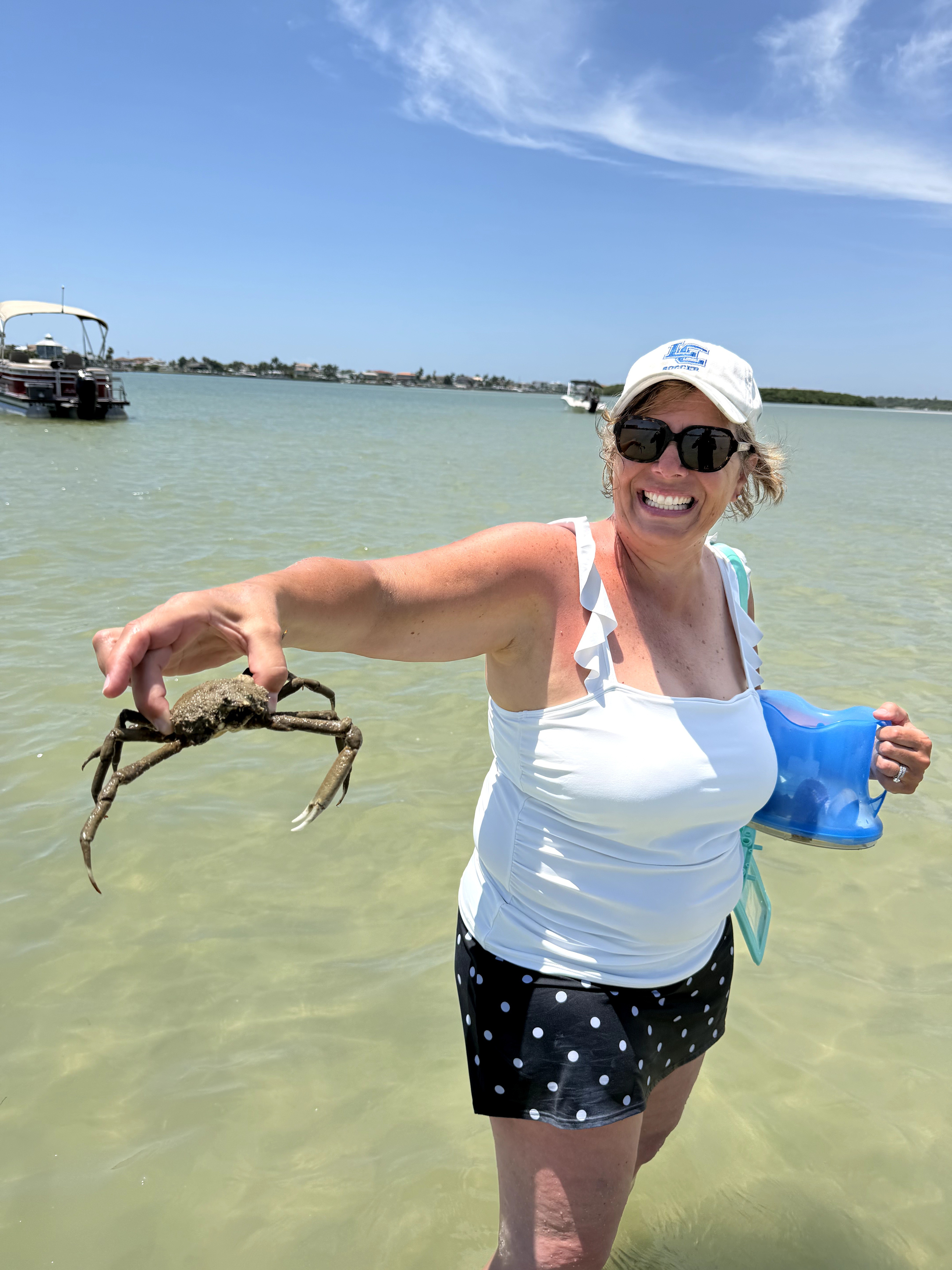 Woman in sunglasses and hat holding a crab in shallow water, smiling, with boats and sky in background.