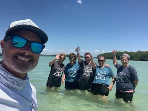 Group of five people standing in water, waving, with a person taking a selfie in the foreground, clear sky, and trees in background.