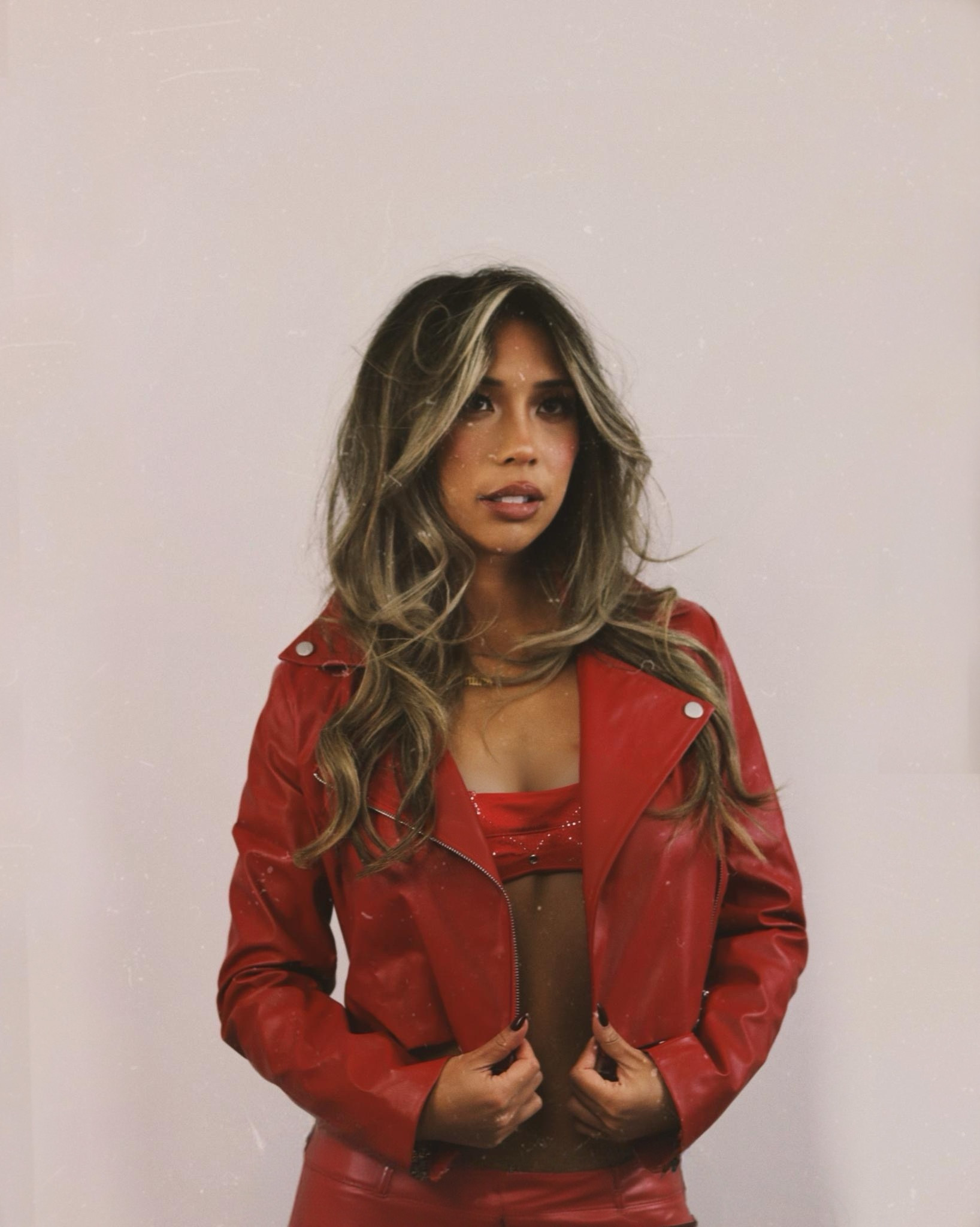 Woman with long wavy hair wearing a red leather jacket and top, standing against a plain background.