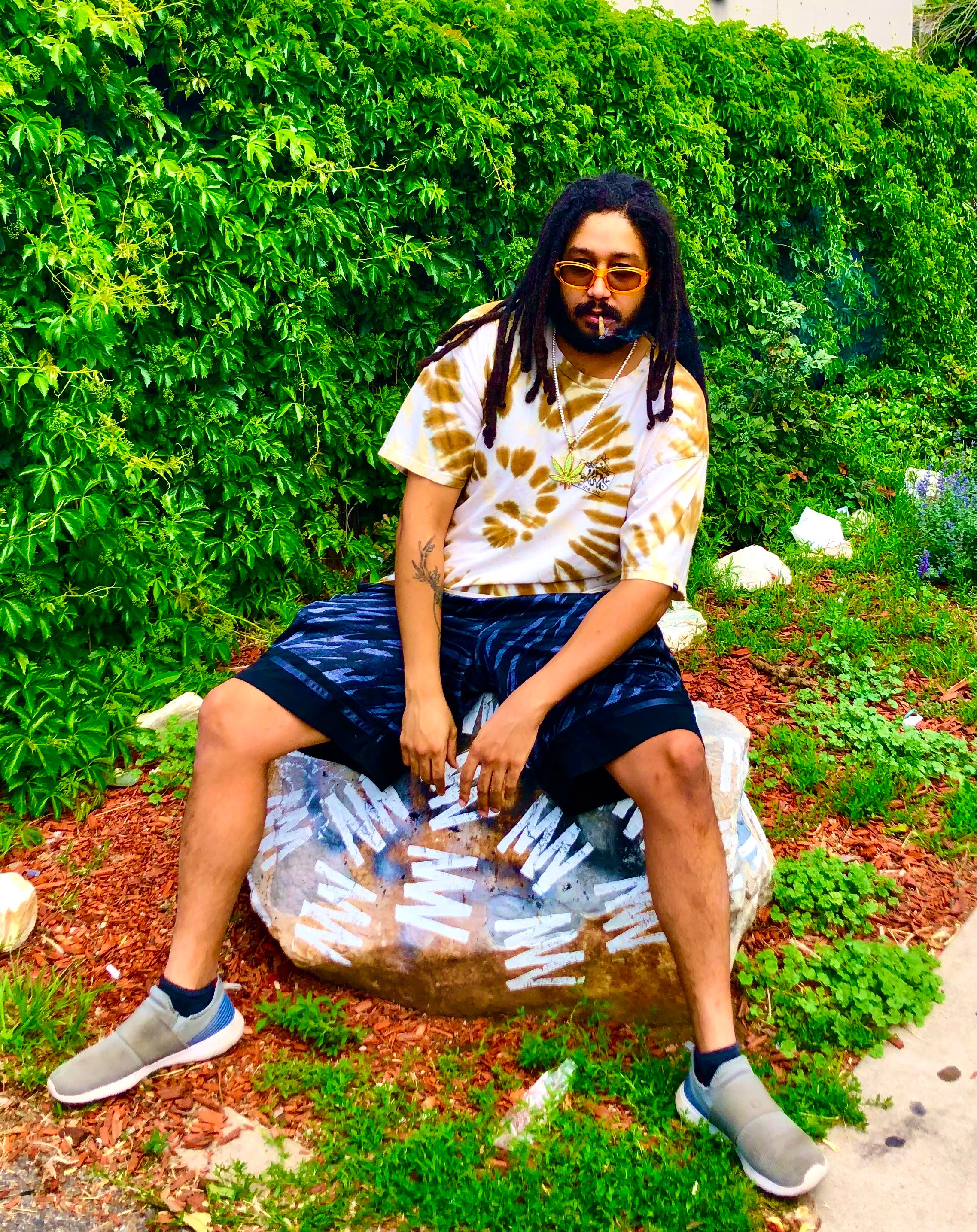 Person with dreadlocks and sunglasses sitting on a plastic bag outdoors, surrounded by green bushes and plants.