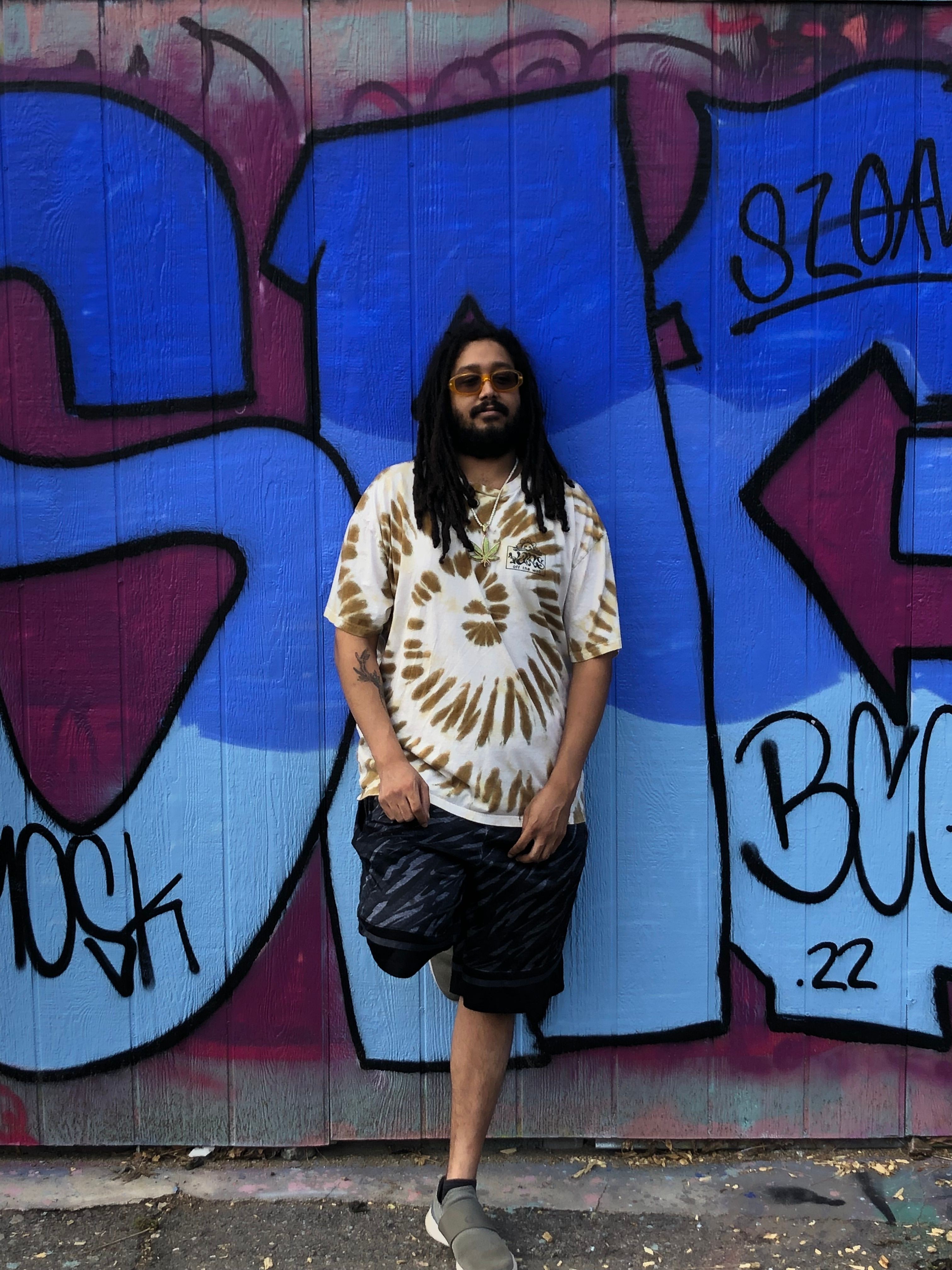 Woman standing in front of colorful graffiti wall, wearing a white and gold patterned top and black skirt, with long dark hair.