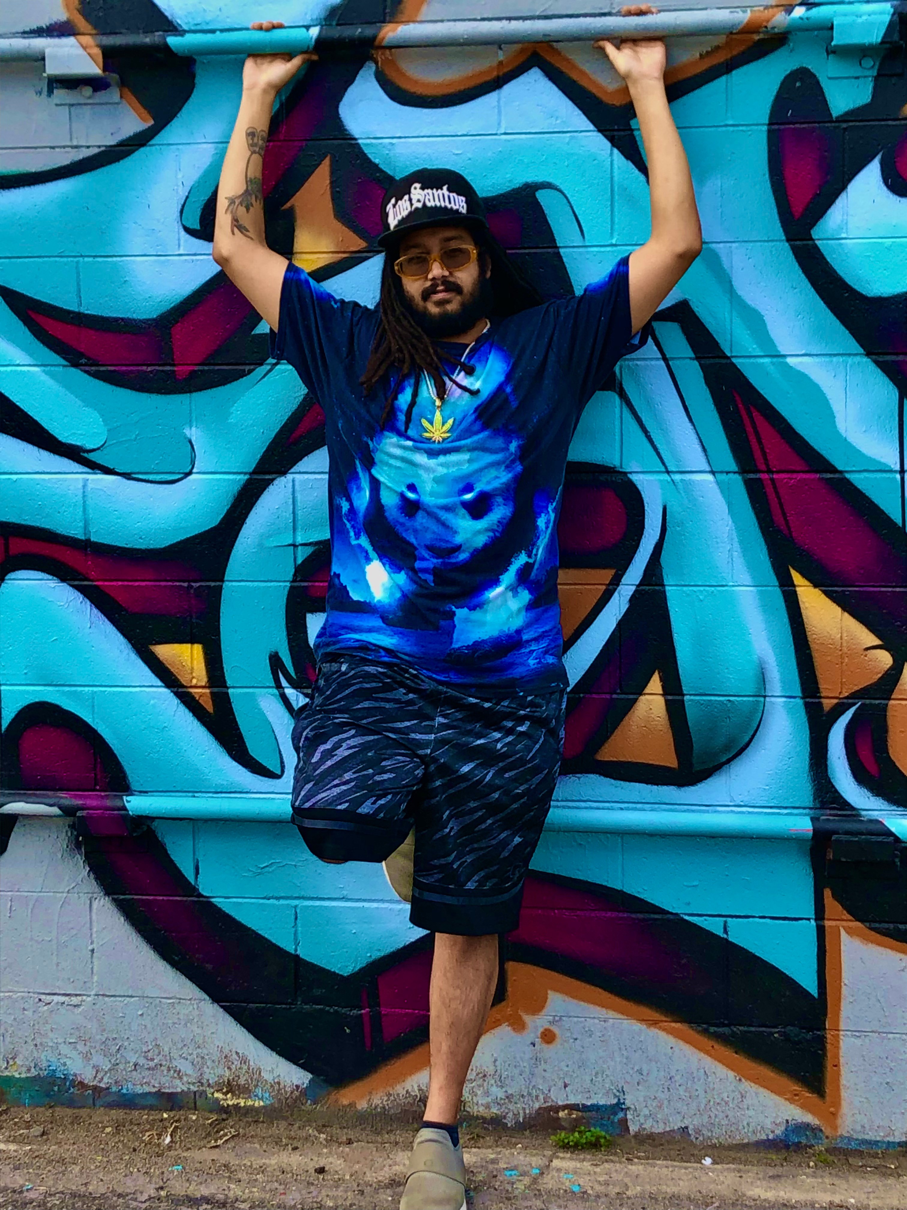 Person with long hair and beard wearing a blue shirt and shorts, standing against a colorful graffiti wall, holding a pipe above head.