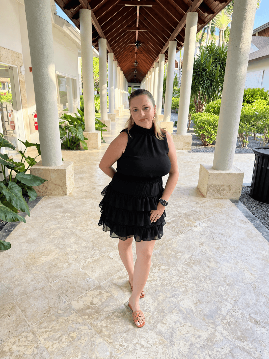 Woman in black dress standing in a covered outdoor walkway with columns and greenery, facing camera, smiling.