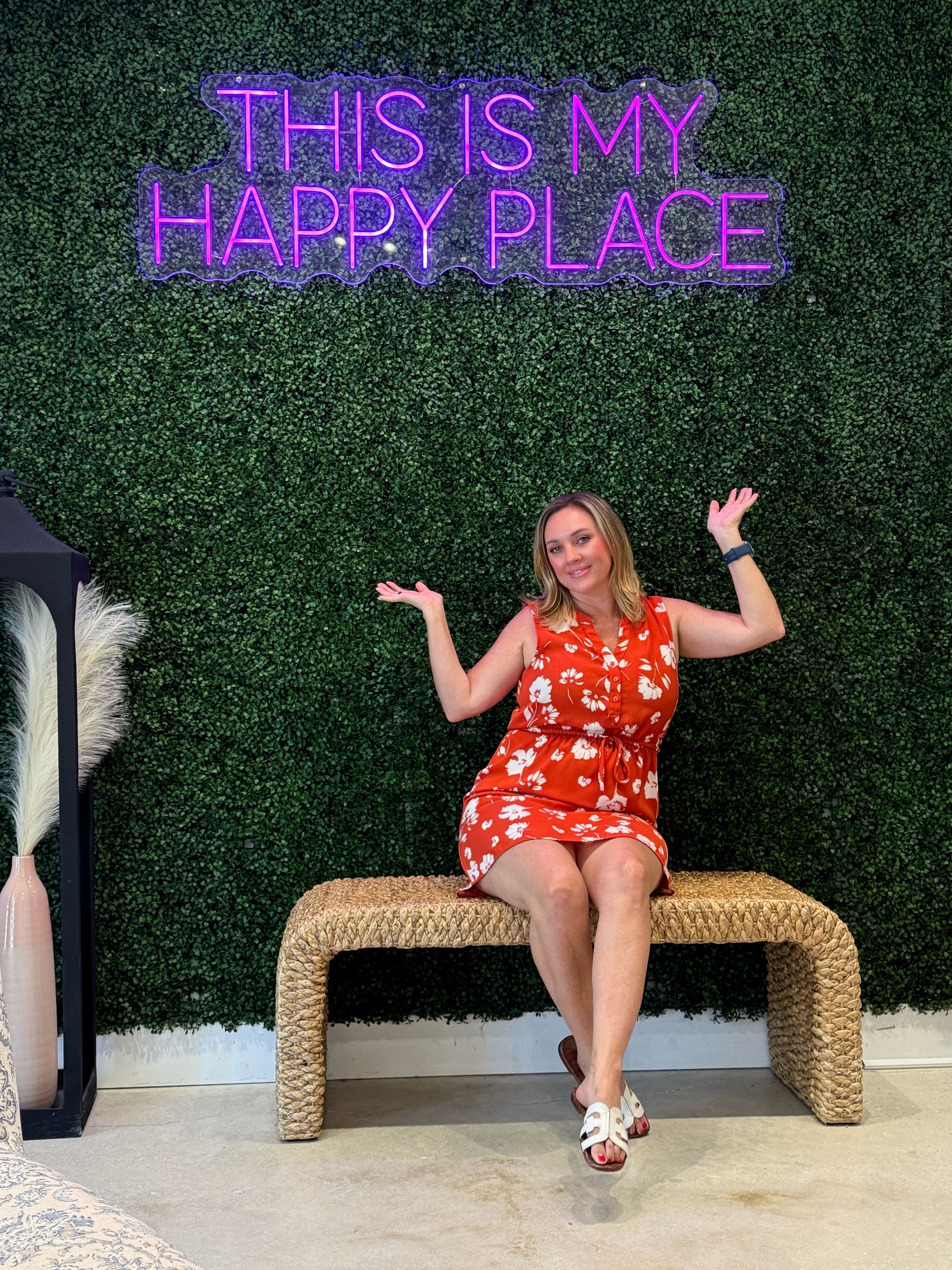 Woman in a red dress sitting on a woven bench, smiling, with a neon sign that reads 'This is my happy place' behind her.