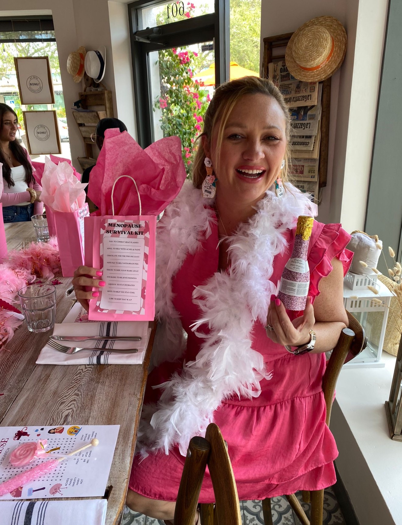 Woman in pink dress with feather boa holding gift bag and bottle, smiling at celebration, with other people and decorations in background.