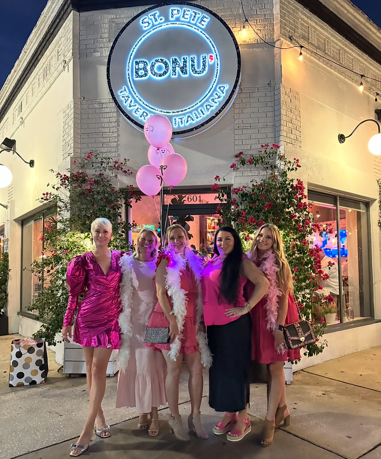 Group of five women standing outside a building with a neon sign reading 'BONUS' and pink balloons, at night.